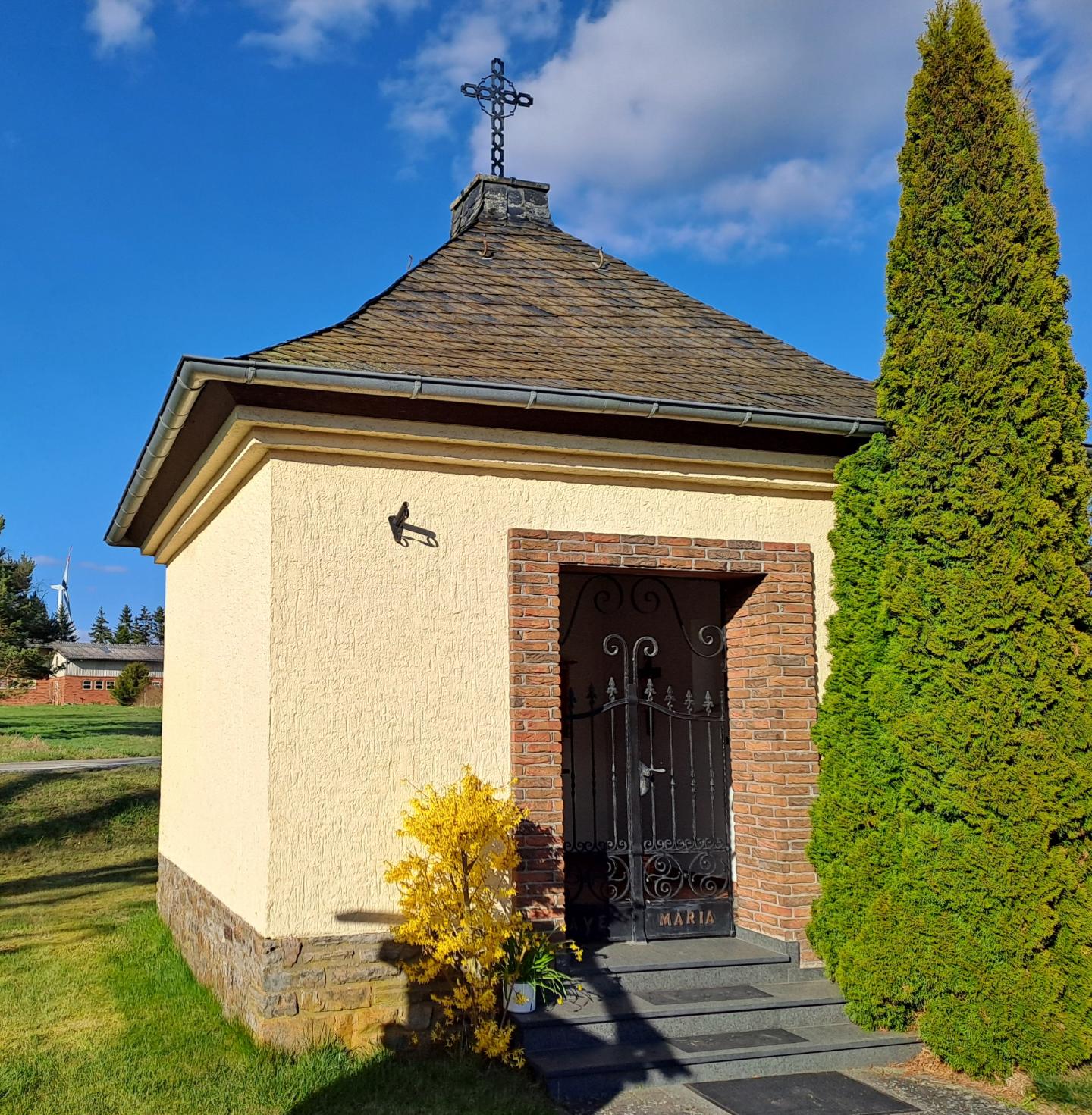 Ein kleines Gebäude mit Metallkreuz auf dem Dach. Rechts davor steht eine Hecke.