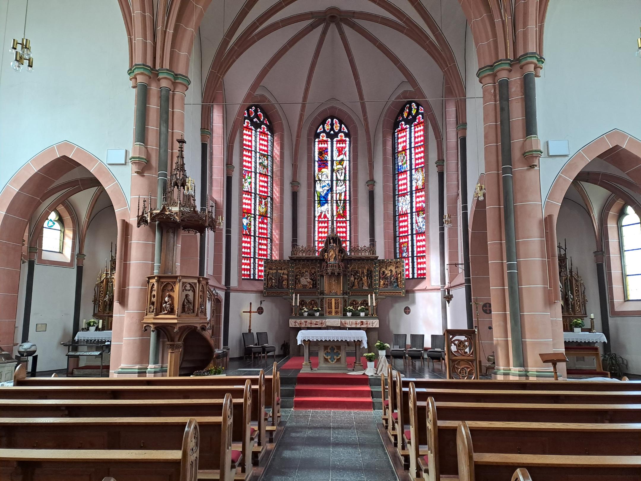 Gesamter Blick auf den Chor mit Hochaltar, bunten Glasfenstern und roten Teppichen. Links ist die hölzerne Kanzel zu sehen, rechts mehrere Seitennischen.