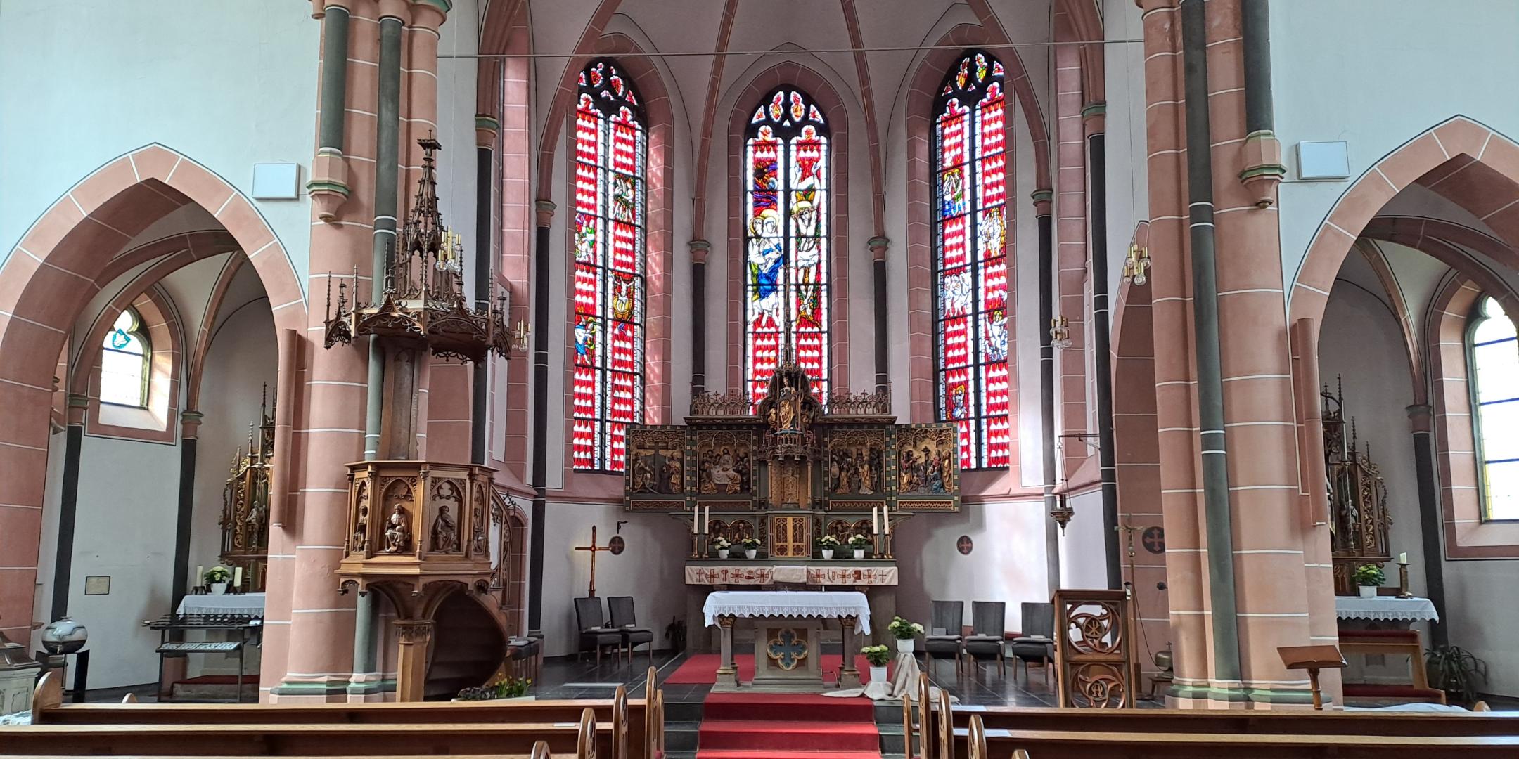 Gesamter Blick auf den Chor mit Hochaltar, bunten Glasfenstern und roten Teppichen. Links ist die hölzerne Kanzel zu sehen, rechts mehrere Seitennischen.