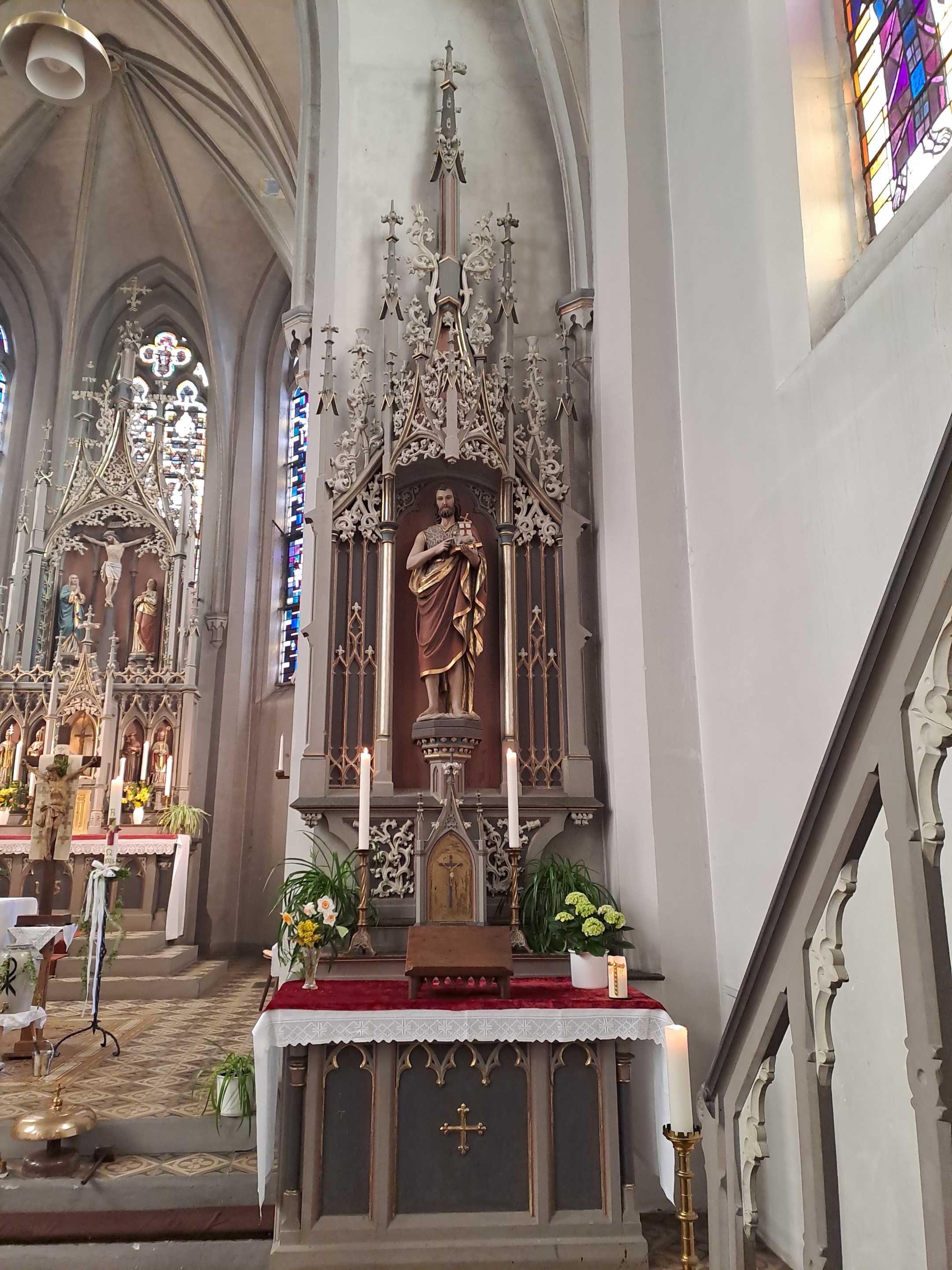 Ein Seitenaltar in einer Kirche, bestehend aus einem grauen Altartisch mit Kerzen und Blumen. Darüber steht eine einzelne, bekleidete Figur des Hl. Johannes des Täufers in einer reich ornamentierten, spitzbogigen Nische aus hellem Stein. Im Hintergrund sind Teile des Hauptaltars sowie farbige Kirchenfenster zu sehen. Rechts führt eine Treppe zur Kanzel.