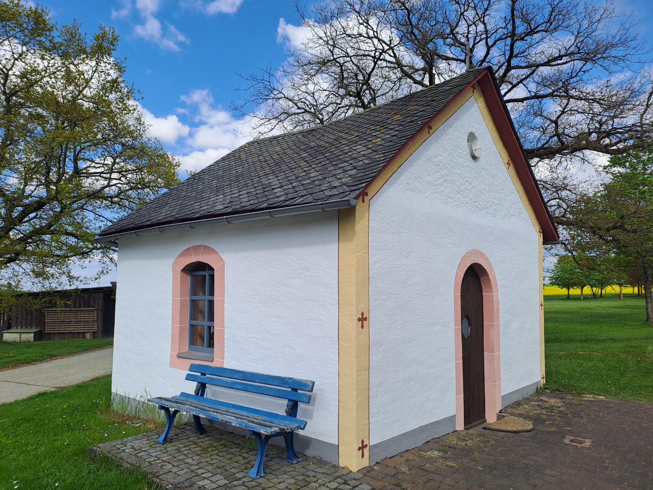 Weiße Kapelle mit kleinem Fenster und Holztür. Vor der Wand steht eine blaue Sitzbank. Die Kapelle ist von Grünfläche und Bäumen umgeben.