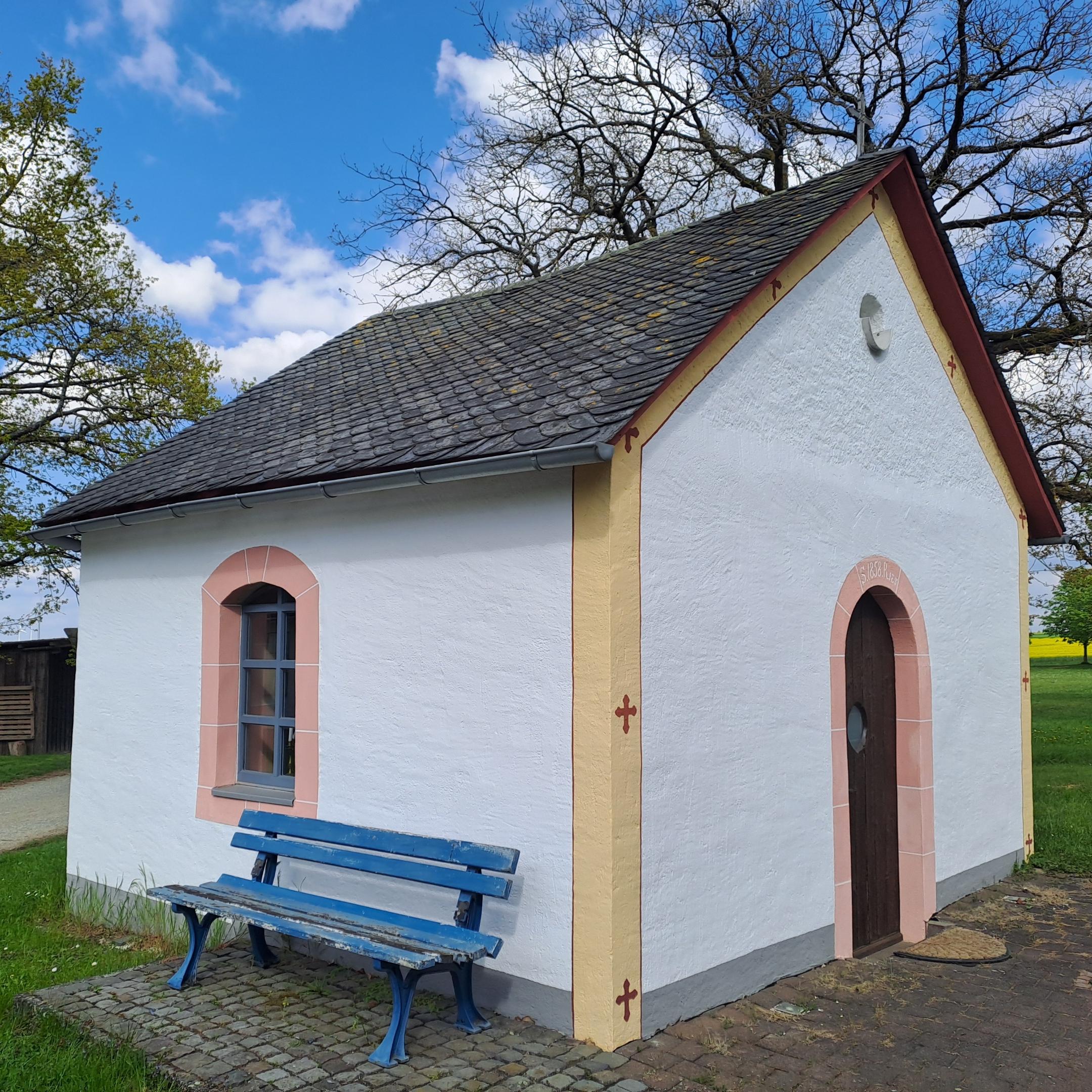 Weiße Kapelle mit kleinem Fenster und Holztür. Vor der Wand steht eine blaue Sitzbank. Die Kapelle ist von Grünfläche und Bäumen umgeben.