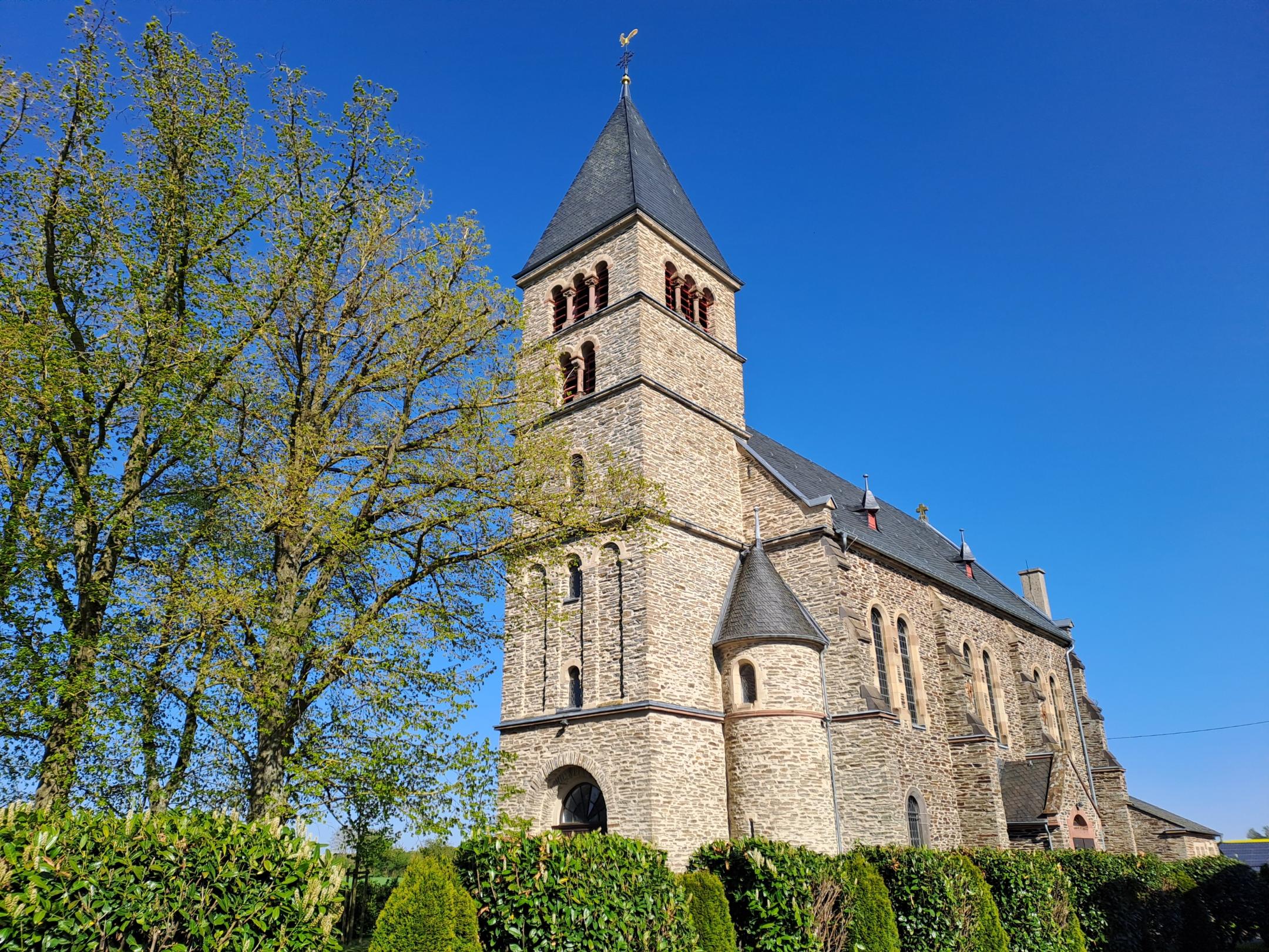Außenansicht einer steinernen Kirche mit hohem Turm und schmalen Fenstern, umgeben von Hecken und Bäumen. Der Himmel ist klar und blau.