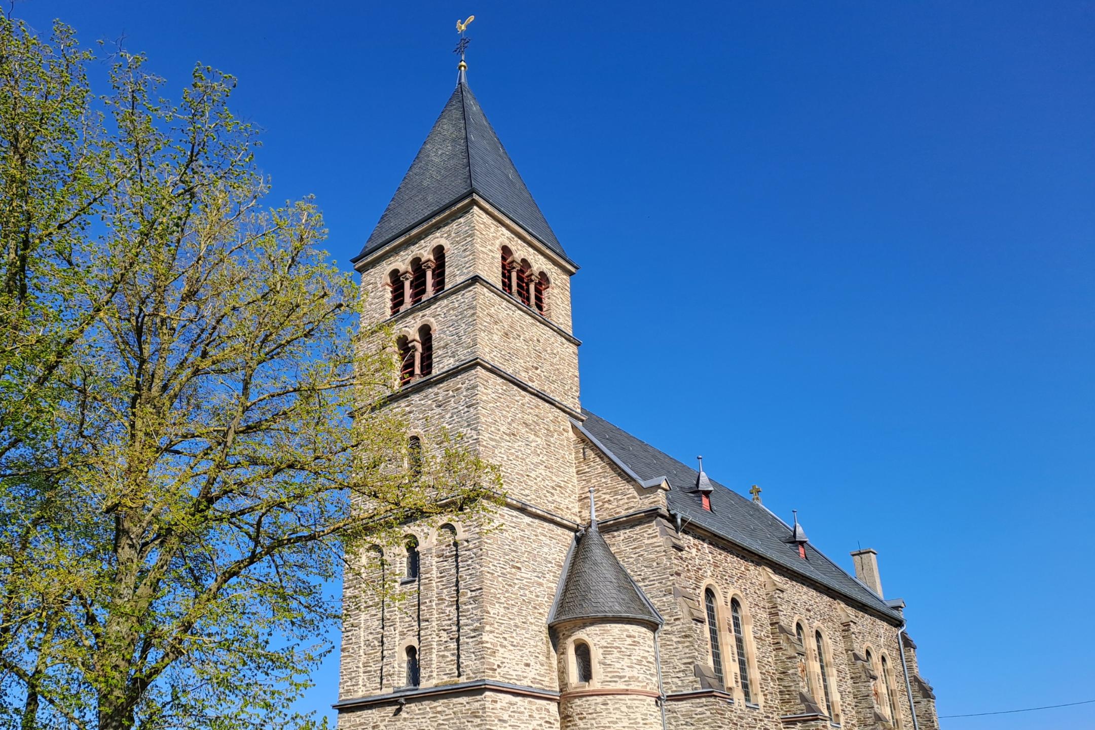 Außenansicht einer steinernen Kirche mit hohem Turm und schmalen Fenstern, umgeben von Hecken und Bäumen. Der Himmel ist klar und blau.