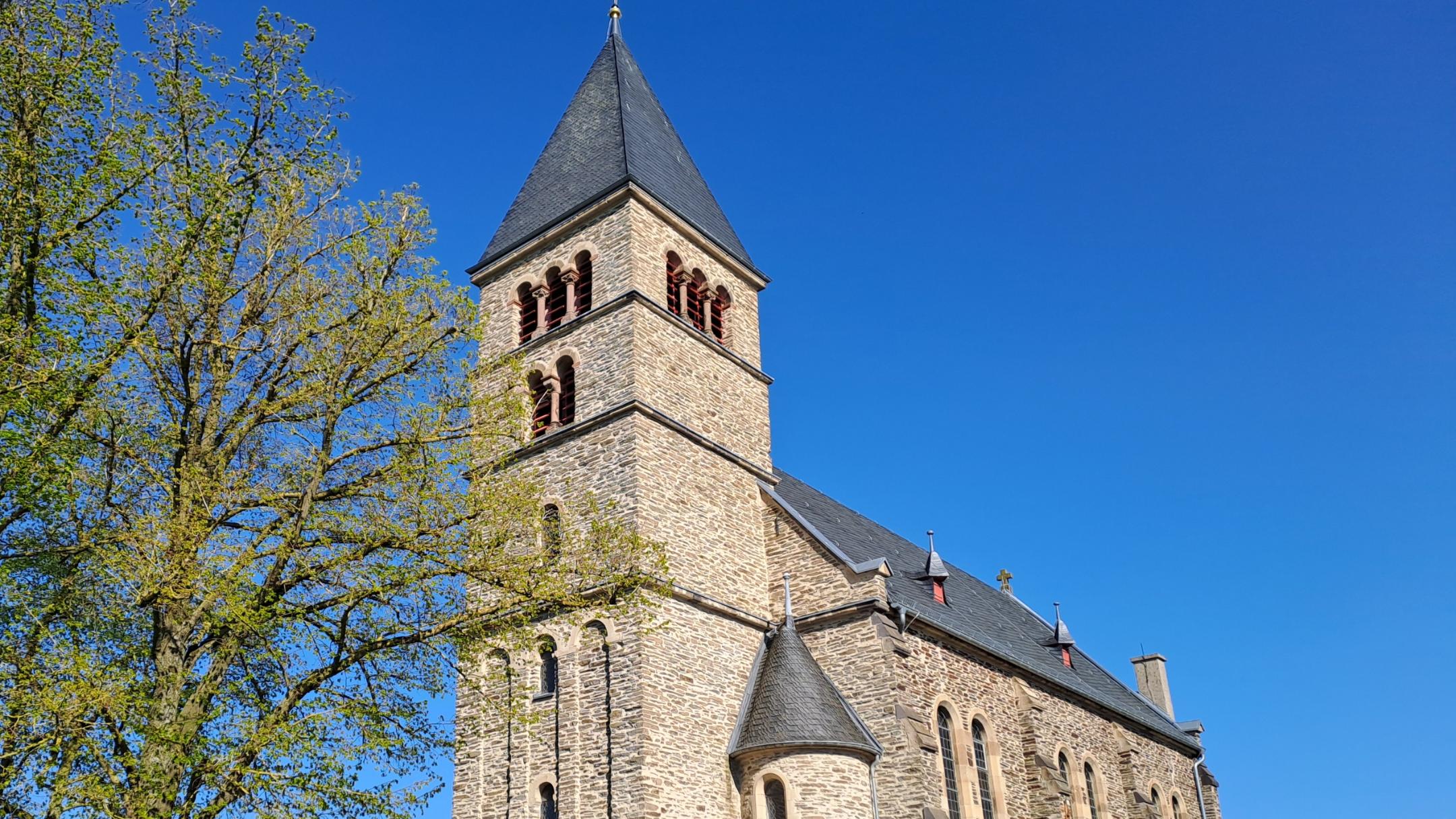 Außenansicht einer steinernen Kirche mit hohem Turm und schmalen Fenstern, umgeben von Hecken und Bäumen. Der Himmel ist klar und blau.