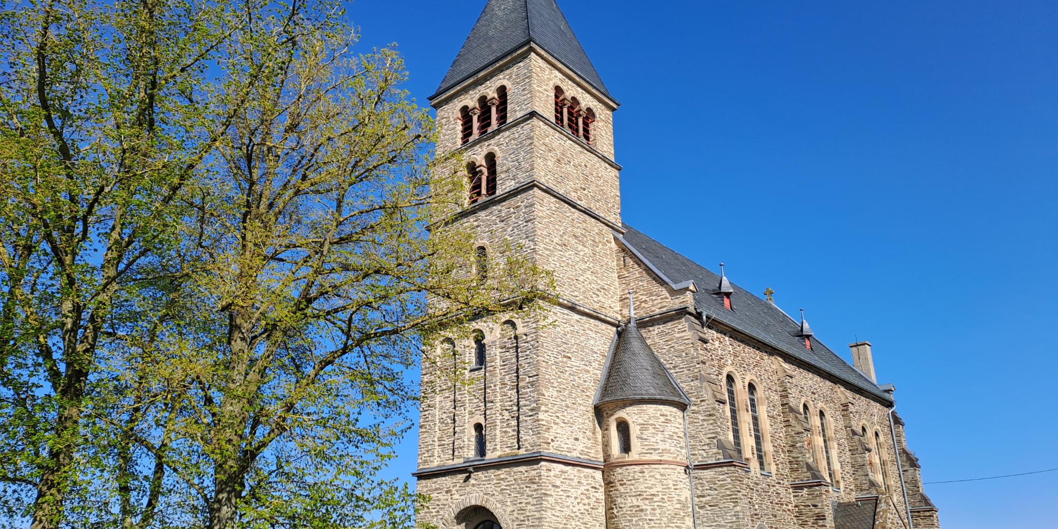 Außenansicht einer steinernen Kirche mit hohem Turm und schmalen Fenstern, umgeben von Hecken und Bäumen. Der Himmel ist klar und blau.