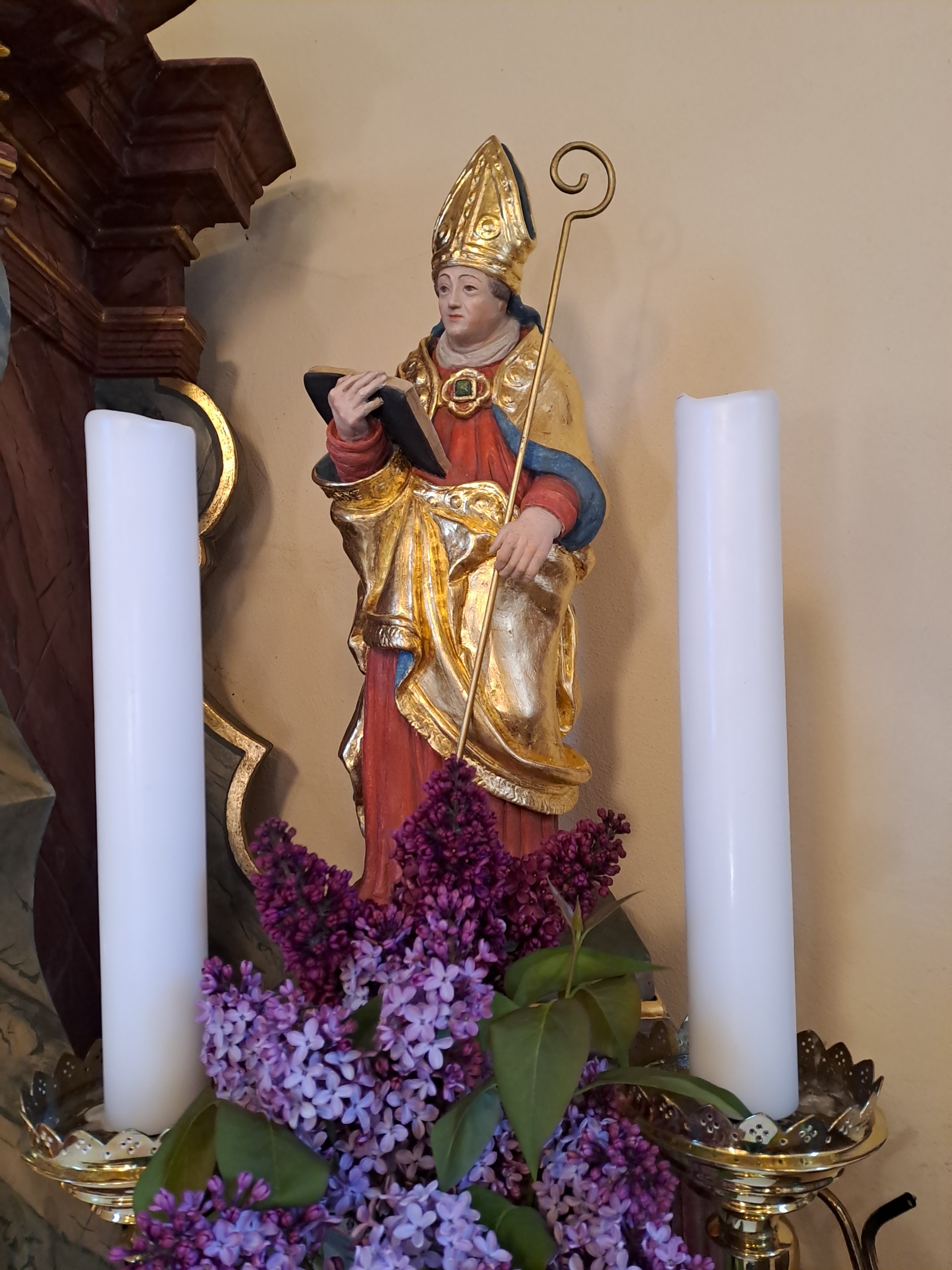 Figur eines Heiligen in goldfarbener Kleidung mit Bischofsstab und Buch, zwischen zwei weißen Kerzen auf dem Altar. Es ist der Hl. Willibrord.