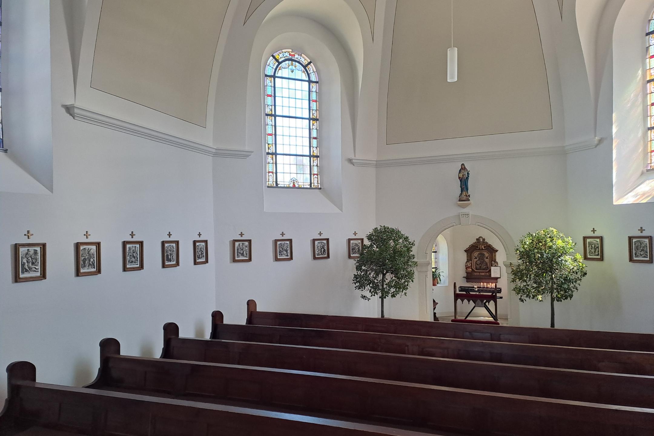 Seitenschiff mit mehreren Holzbänken und einer Reihe kleiner gerahmter Kreuzwegstationen an der weißen Wand. Am Ende des Raums steht eine kleine Kapelle mit Marienbild.