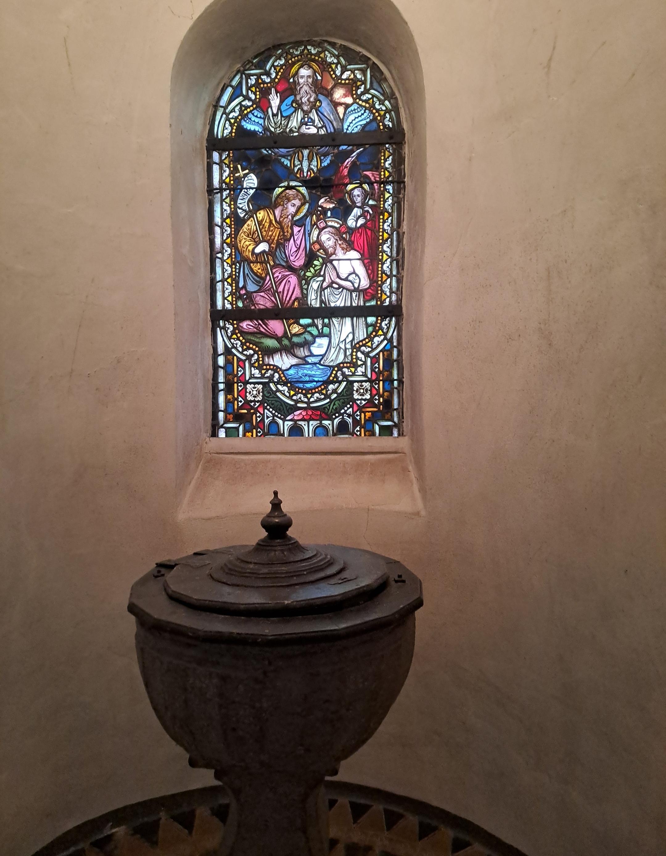 Steinerner Taufstein mit rundem Becken im Vordergrund. Dahinter ein kleines Rundbogenfenster mit Buntglasdarstellung einer religiösen Szene. Die Wände des Raumes sind schlicht und hell gehalten. (c) Christian Jager Steinerner Taufstein mit rundem Becken im Vordergrund. Dahinter ein kleines Rundbogenfenster mit Buntglasdarstellung einer religiösen Szene. Die Wände des Raumes sind schlicht und hell gehalten.
