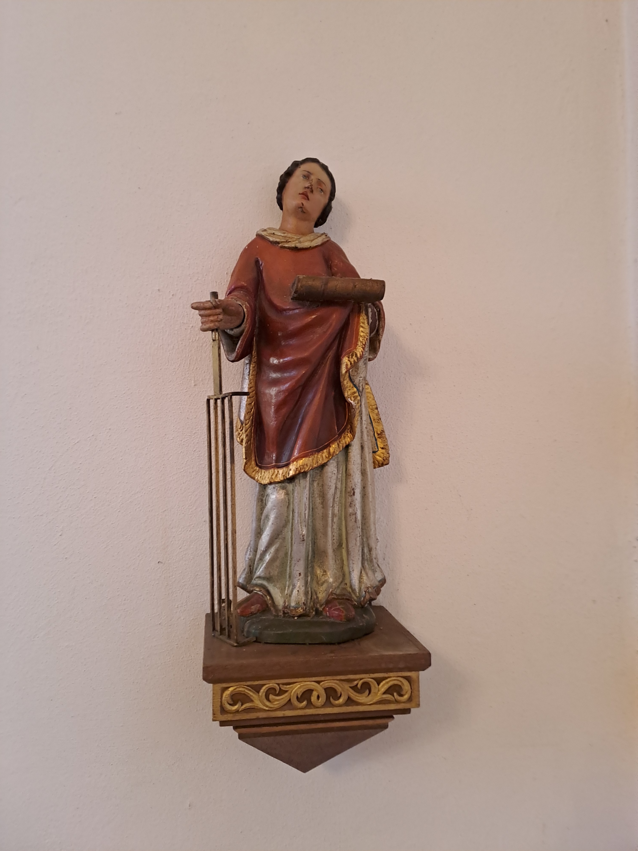 Holzfigur an der Kirchenwand auf einem kleinen Wandpodest. Die Figur hält ein Buch in einer Hand und ein rostartiges Attribut in der anderen. Die Kleidung ist farbig gefasst, der Hintergrund ist eine helle Wand.