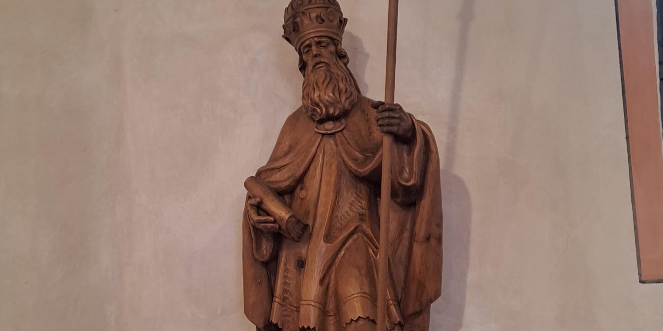 Geschnitzte Holzfigur eines bärtigen Heiligen oder Bischofs mit Mitra und langem Stab. Die Figur steht auf einer Wandkonsole. Zu Füßen der Skulptur befindet sich eine kleine Blumenvase. Im Hintergrund ist eine helle Kirchenwand zu sehen.
