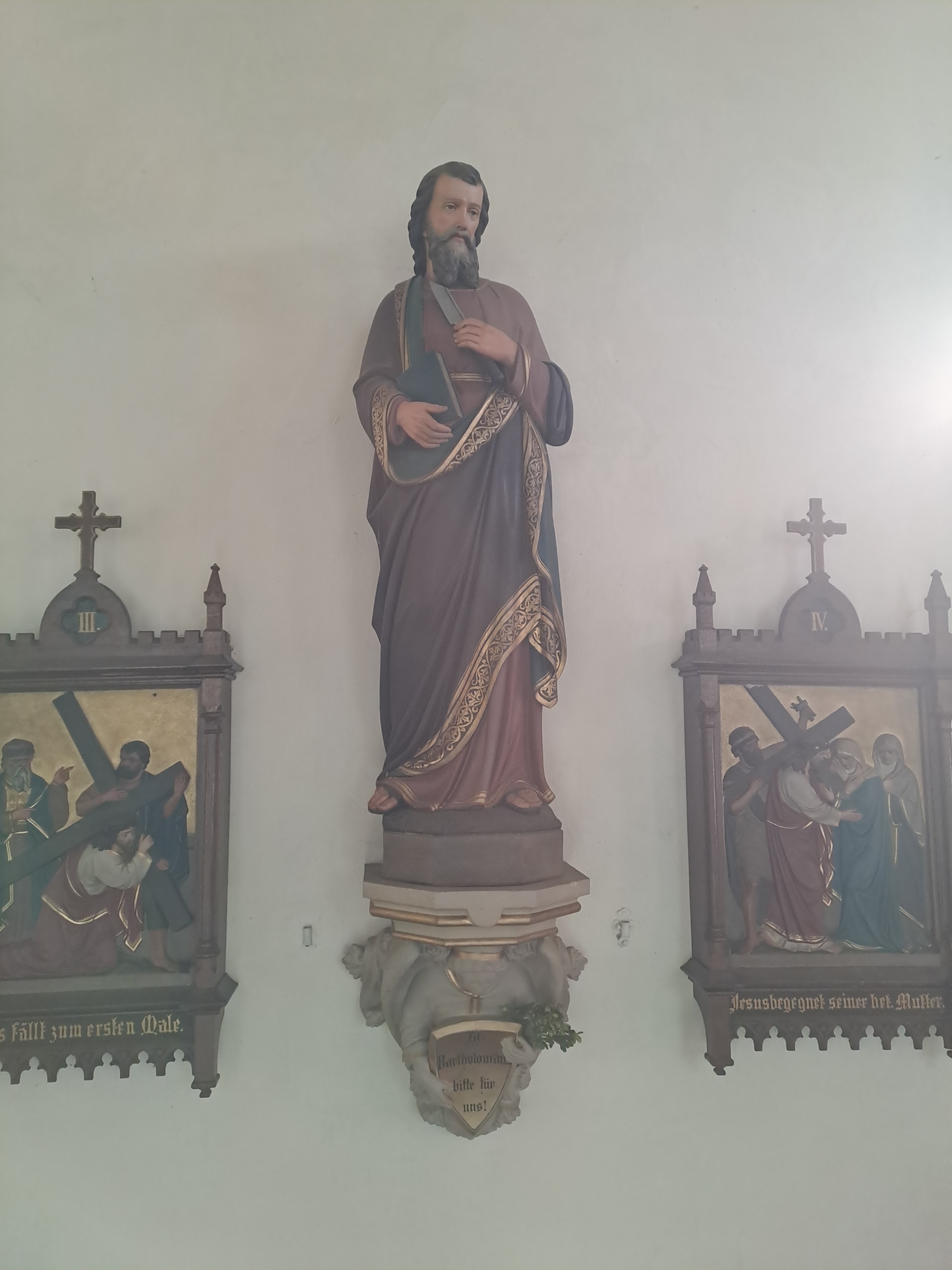 Statue eines Heiligen mit Buch, umgeben von Kreuzwegdarstellungen.