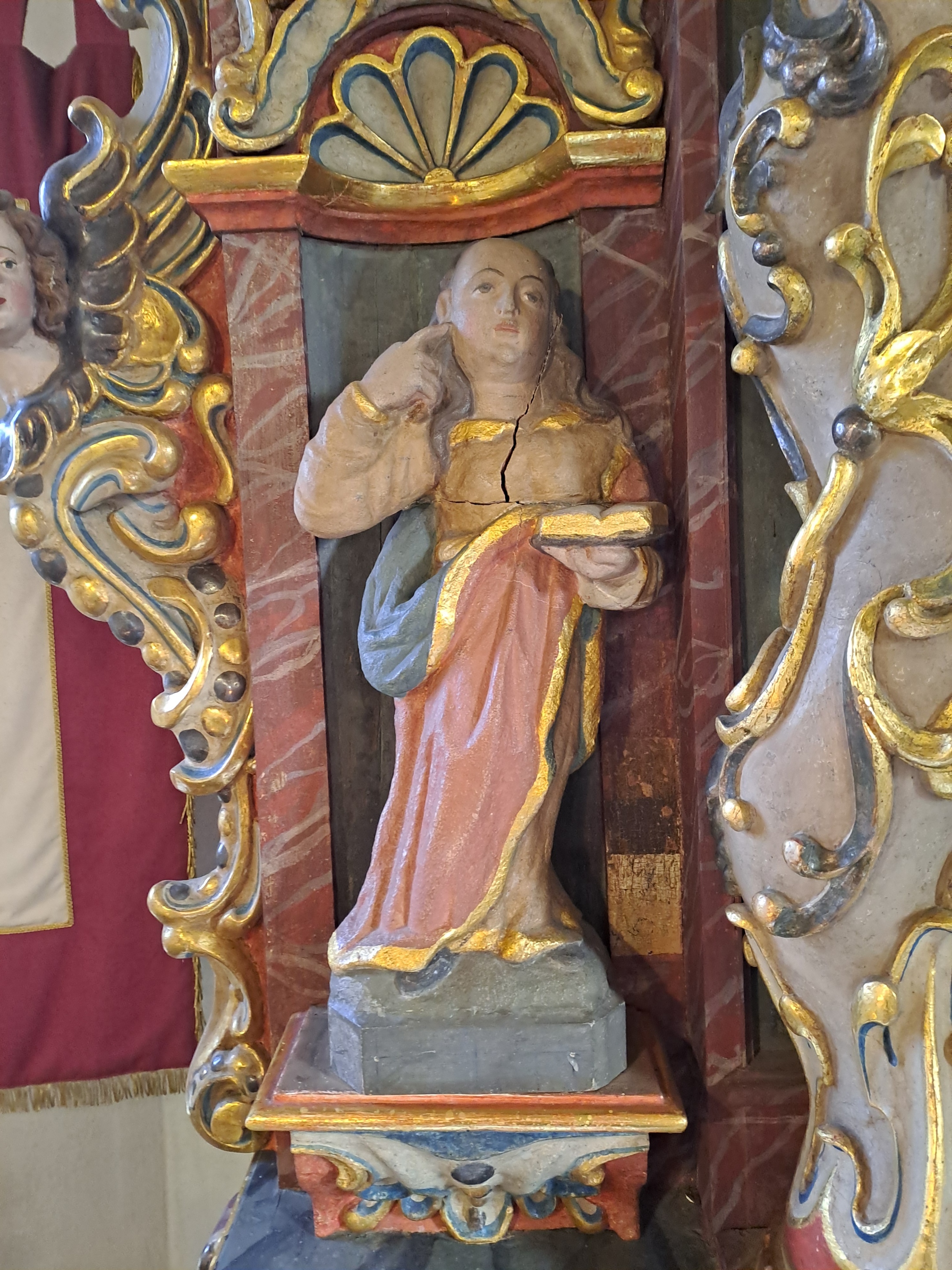 Detail einer bemalten Heiligenfigur am Altar mit Buch in der Hand und sichtbarer Rissspur im Material.