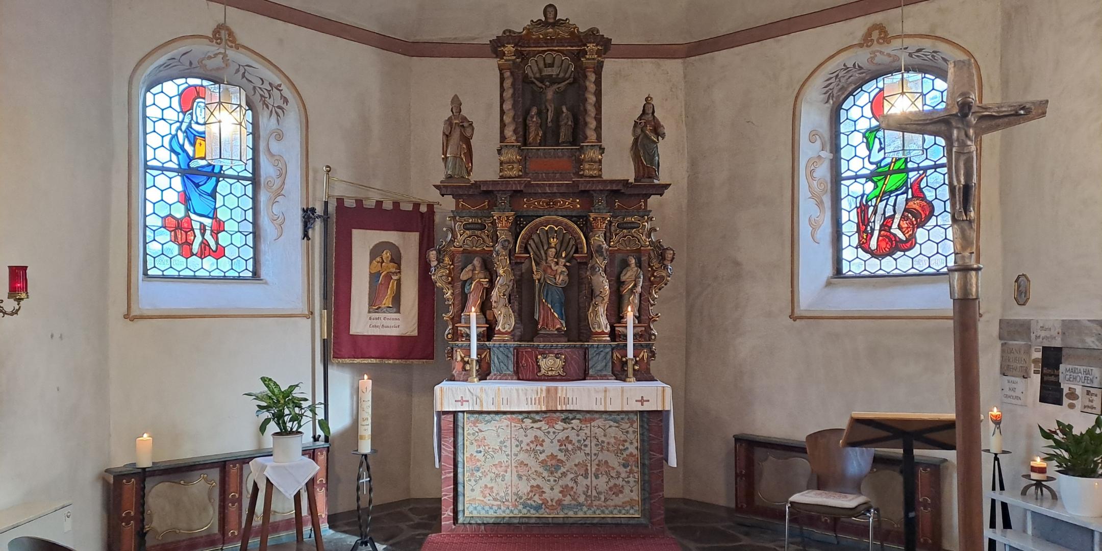 Innenraum einer kleinen Kirche mit zentralem Altar, Buntglasfenstern und freistehendem Kreuz im Vordergrund.