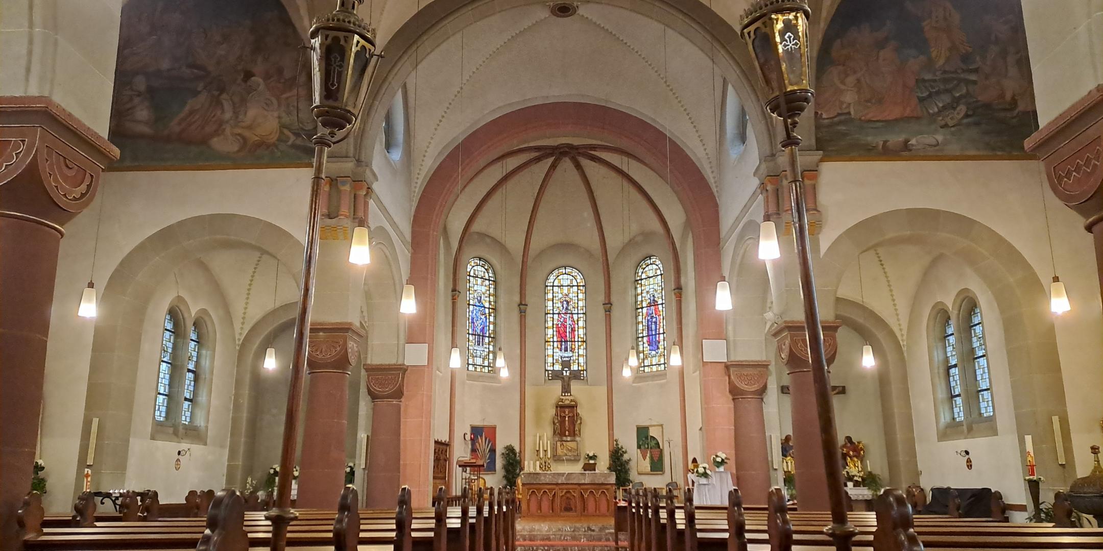 Blick durch den Mittelgang einer Kirche mit Holzbänken auf beiden Seiten. Ein roter Teppich führt zum Altar im Hintergrund. Über dem Altar befinden sich drei bunte Glasfenster und ein Kreuz. Säulen und Rundbögen tragen das Gewölbe, an der Decke hängen Leuchten.