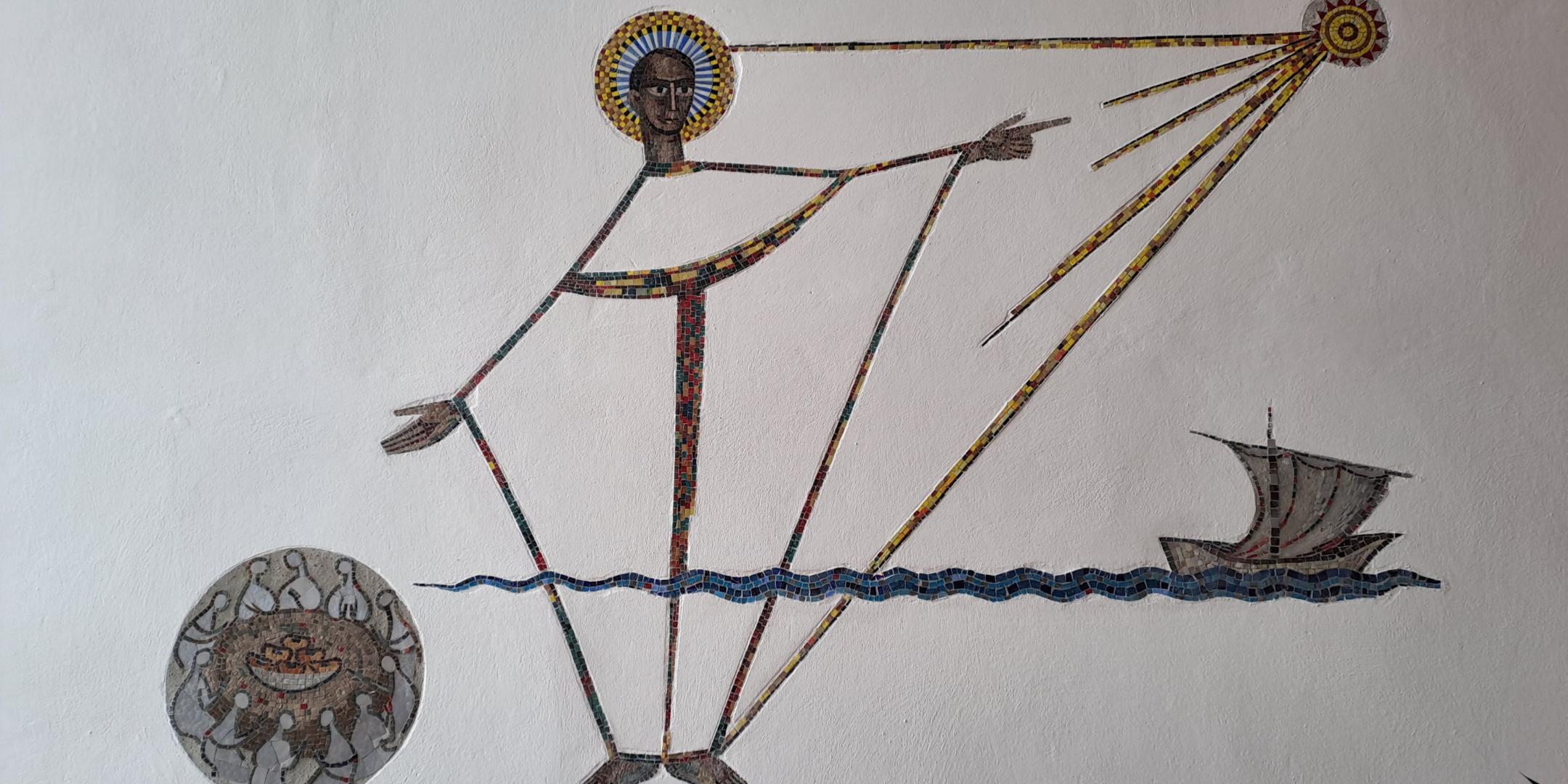 Wandmosaik in einer Kirche mit einer stilisierten menschlichen Figur aus kleinen bunten Steinen. Die Figur steht im Wasser und zeigt mit ausgestrecktem Arm auf eine kreisförmige Sonne, von der Strahlen ausgehen. Rechts ist ein kleines Boot mit Segel dargestellt, links ein rundes Symbol mit dekorativen Mustern. Der Hintergrund ist eine helle, glatte Wand.