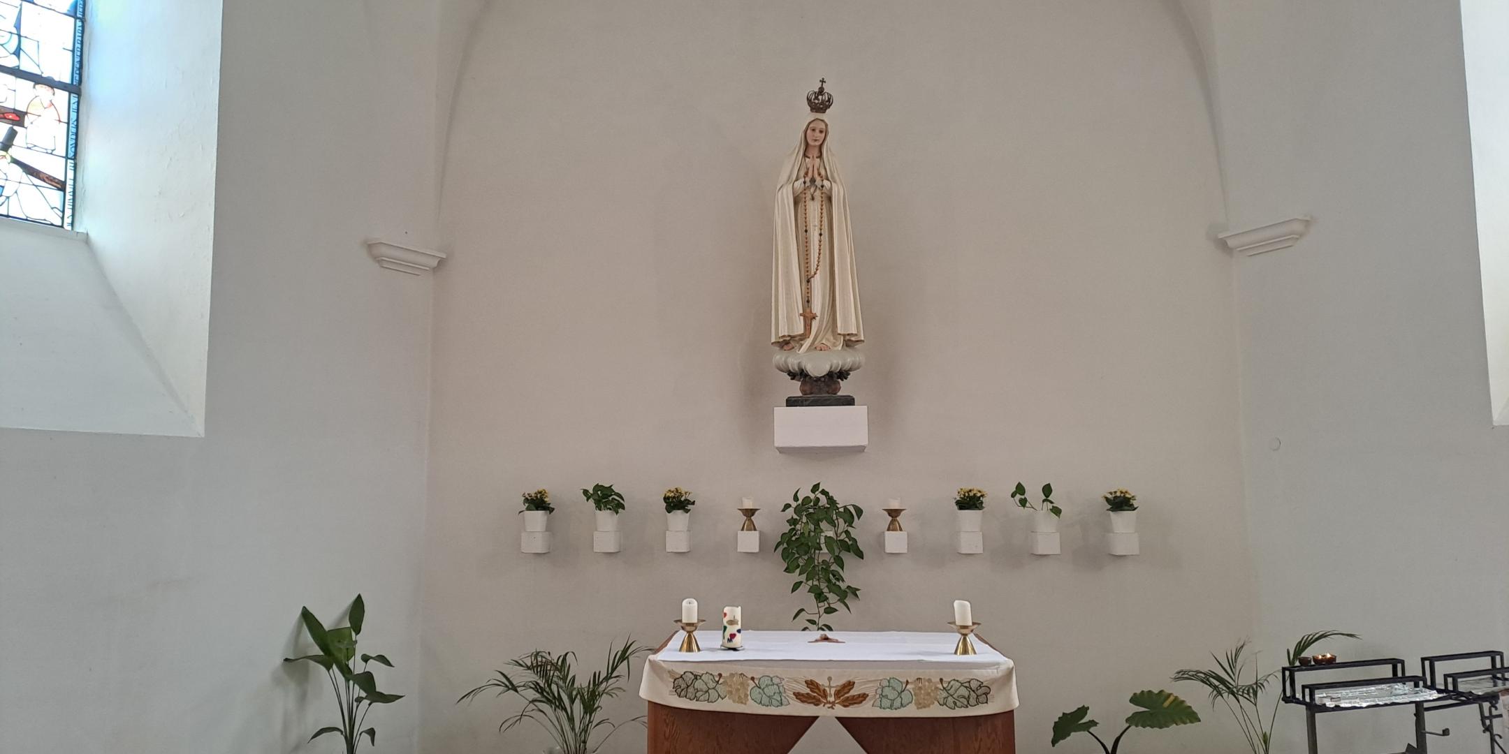 Seitenaltar in einer Kirche mit einer Statue auf einem Wandpodest. Die Figur trägt ein langes Gewand, eine Krone und hält einen Rosenkranz. Unterhalb befindet sich ein schlichter Altar mit Kerzen und einer Altardecke. Entlang der Wand stehen mehrere kleine Pflanzen in weißen Töpfen. Die Wand ist hell und halbrund ausgeführt.