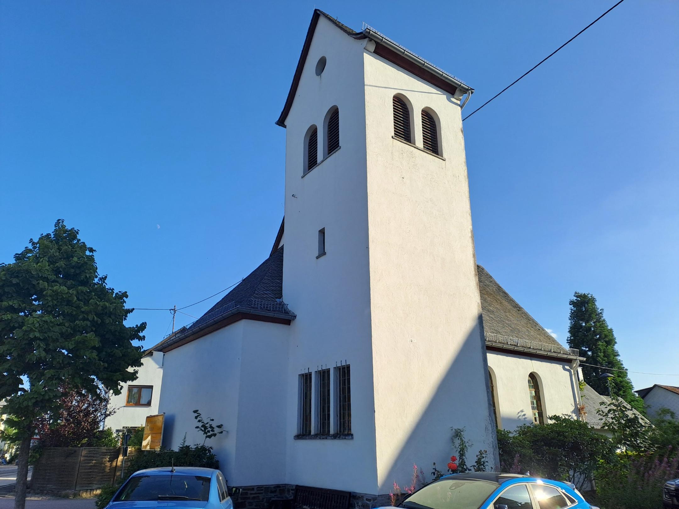 Weiße Kirche mit rechteckigem Glockenturm unter blauem Himmel, umgeben von Bäumen und Wohnhäusern.
