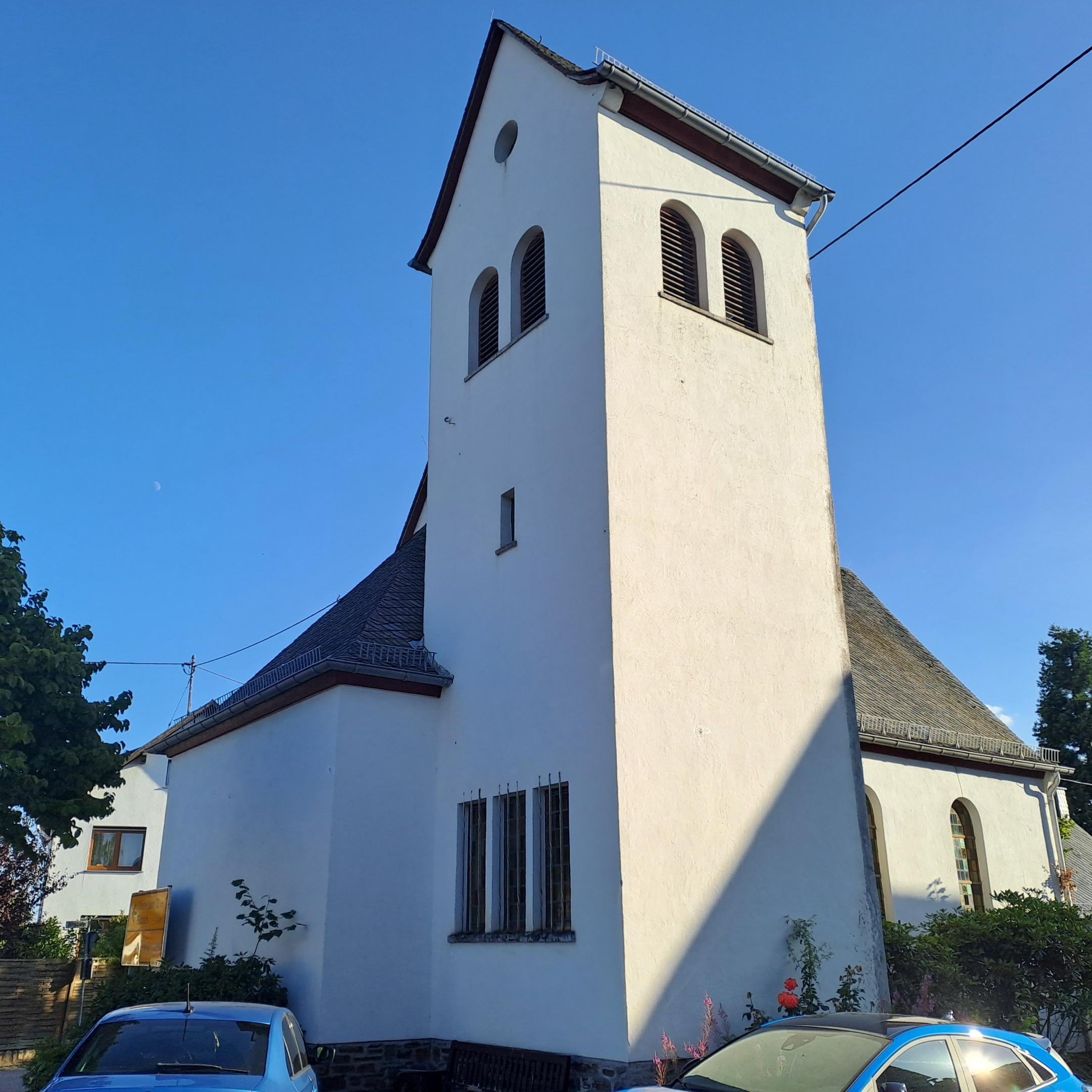 Weiße Kirche mit rechteckigem Glockenturm unter blauem Himmel, umgeben von Bäumen und Wohnhäusern.