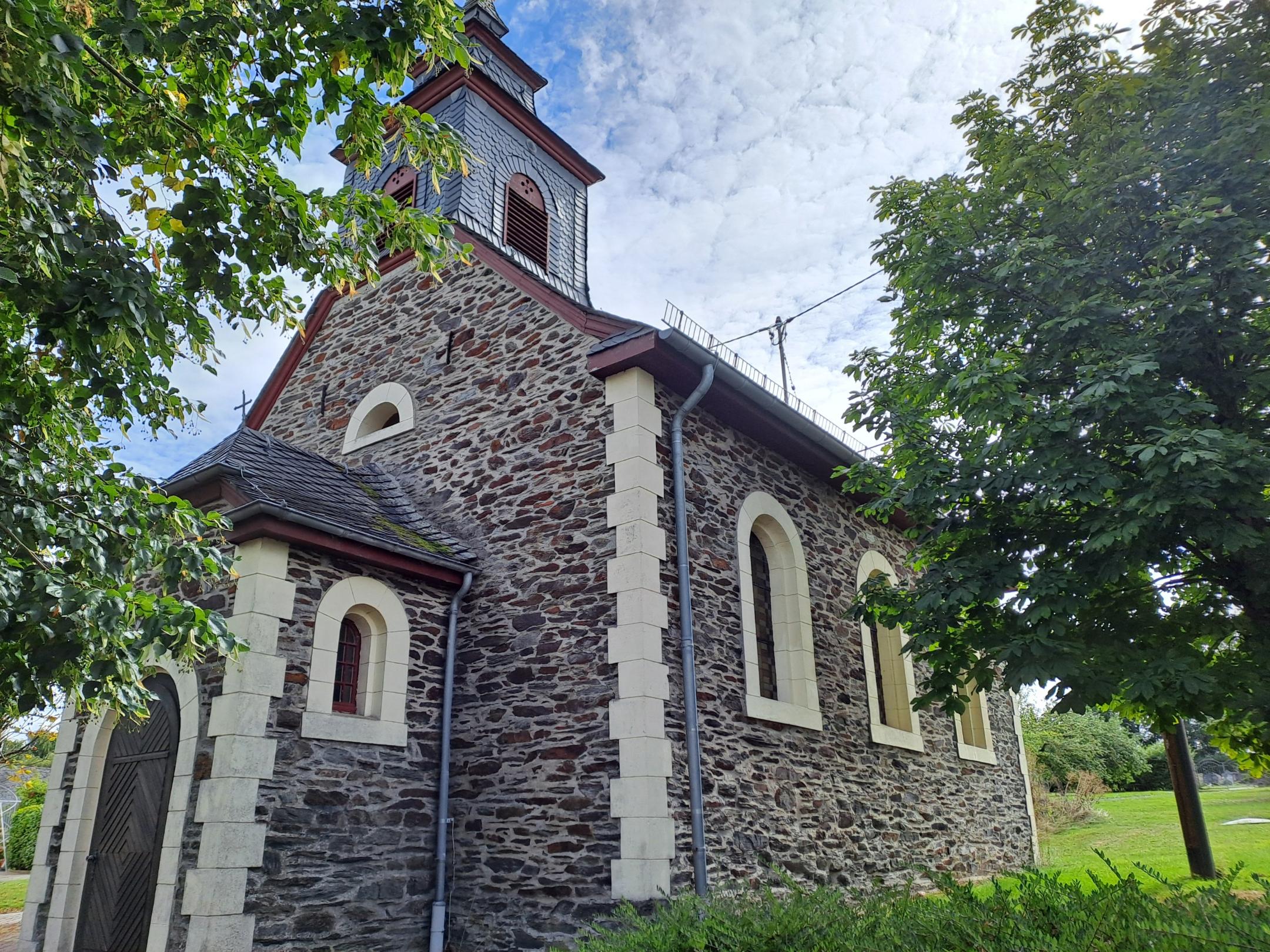 Seitenansicht der Kirche St. Josef in Alterkülz. Das Bruchsteingebäude mit Rundbogenfenstern und Dachreiter ist von Bäumen und Grünflächen umgeben.