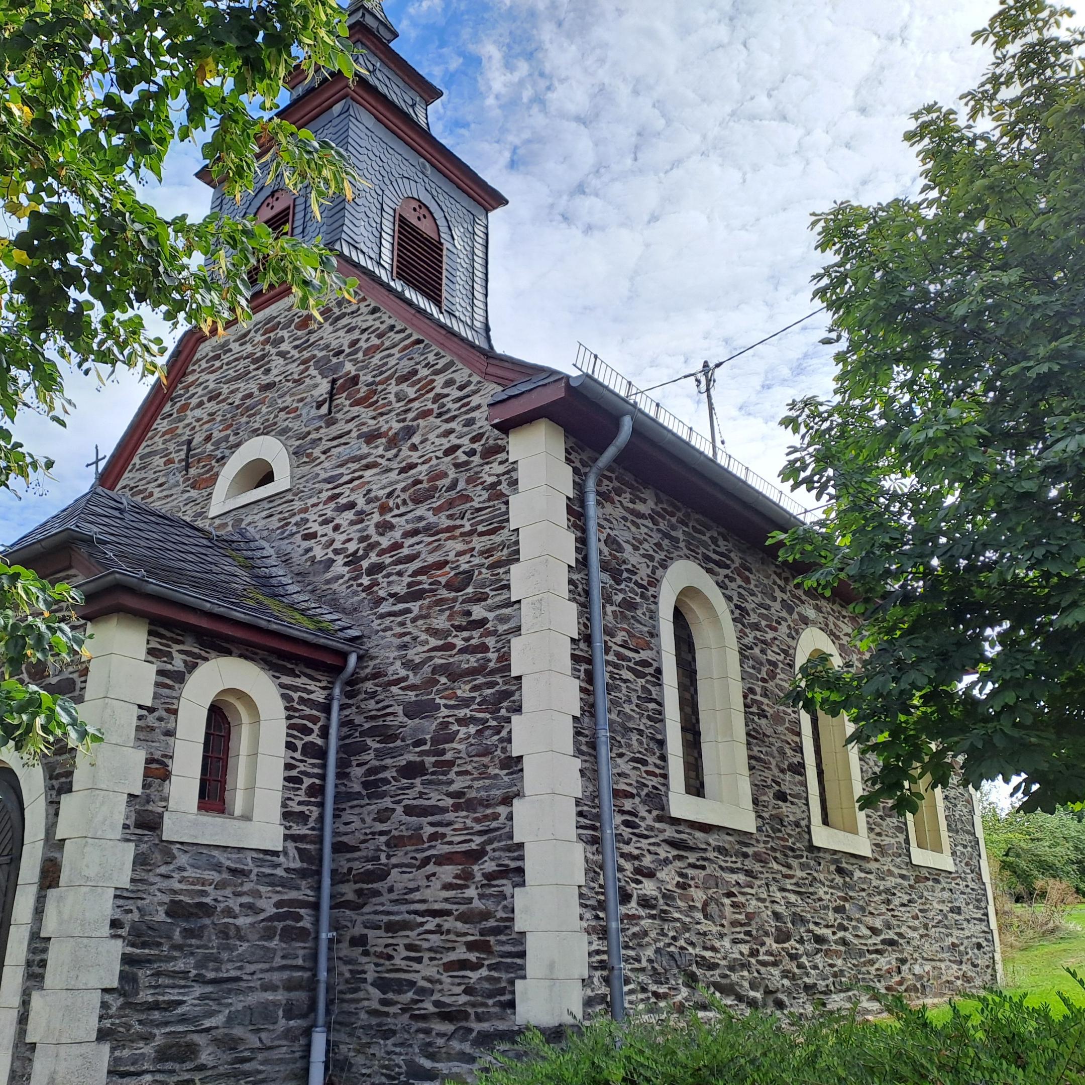 Seitenansicht der Kirche St. Josef in Alterkülz. Das Bruchsteingebäude mit Rundbogenfenstern und Dachreiter ist von Bäumen und Grünflächen umgeben.