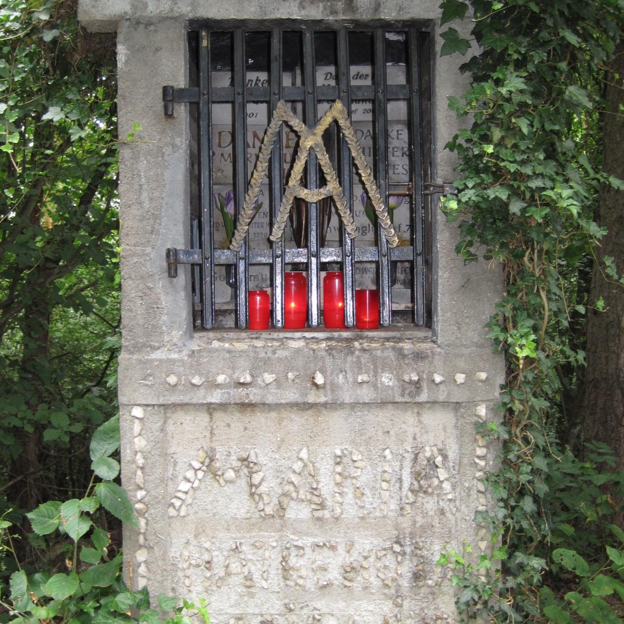 Ein gemauerter Bildstock aus hellem Stein steht am Rand eines bewaldeten Weges. Die Ädikula ist von Efeu überwachsen und durch ein schwarzes Metallgitter verschlossen. Hinter dem Gitter sind Gedenktafeln zu erkennen. Am unteren Rand stehen mehrere rote Grablichter.