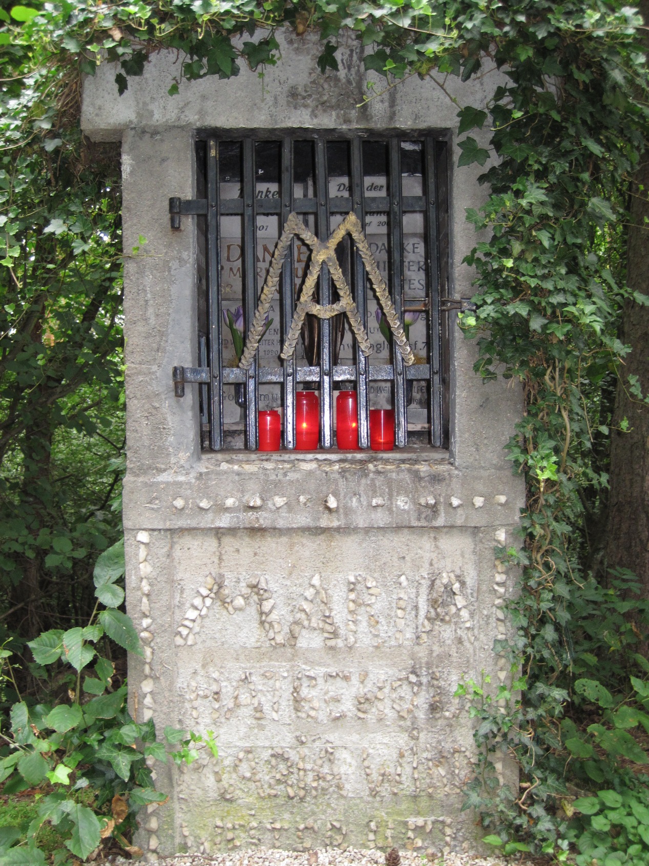 Ein gemauerter Bildstock aus hellem Stein steht am Rand eines bewaldeten Weges. Die Ädikula ist von Efeu überwachsen und durch ein schwarzes Metallgitter verschlossen. Hinter dem Gitter sind Gedenktafeln zu erkennen. Am unteren Rand stehen mehrere rote Grablichter.