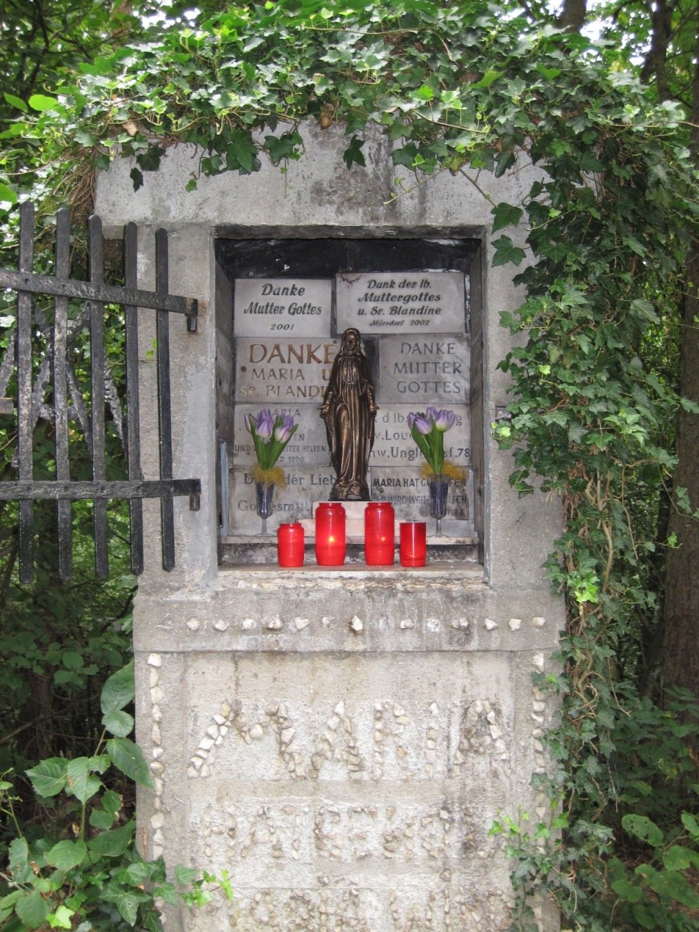 Ein steinerner Bildstock im Wald mit geöffneter schwarzer Gittertür. In einer Nische steht eine kleine Marienfigur aus Bronze. Daneben stecken zwei Blumen in Vasen. Vor der Figur stehen mehrere rote Grablichter. Im Hintergrund sind beschriftete Gedenktafeln angebracht.