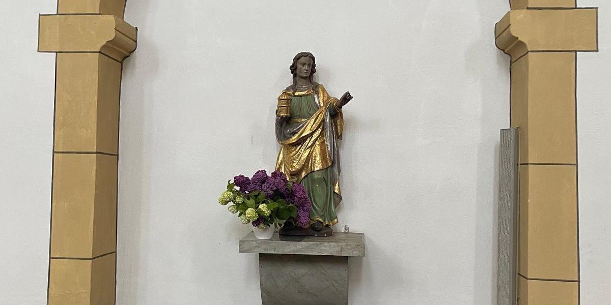 Eine geschnitzte Figur steht in einer kleinen Wandnische unter einem sandfarbenen Steinbogen. Die Figur trägt ein langes Gewand mit goldfarbenen Elementen. Darunter befindet sich ein kleiner steinerner Sockel mit einem Blumenstrauß. Seitlich sind Kirchenbänke zu sehen.