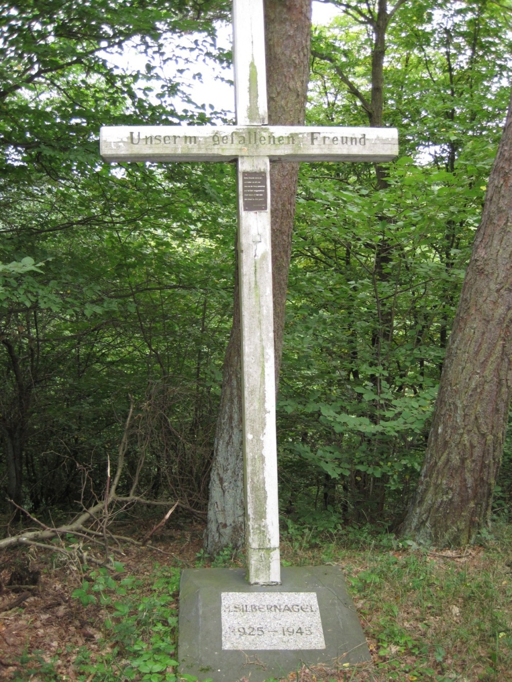 Ein schlichtes, helles Steinkreuz steht in einem Waldgebiet. Am Querbalken ist die Inschrift „Unserem gefallenen Freund“ eingraviert. Am Sockel befindet sich eine weitere beschriftete Steinplatte mit Jahreszahlen. Das Kreuz ist von Bäumen und dichtem Grün umgeben.