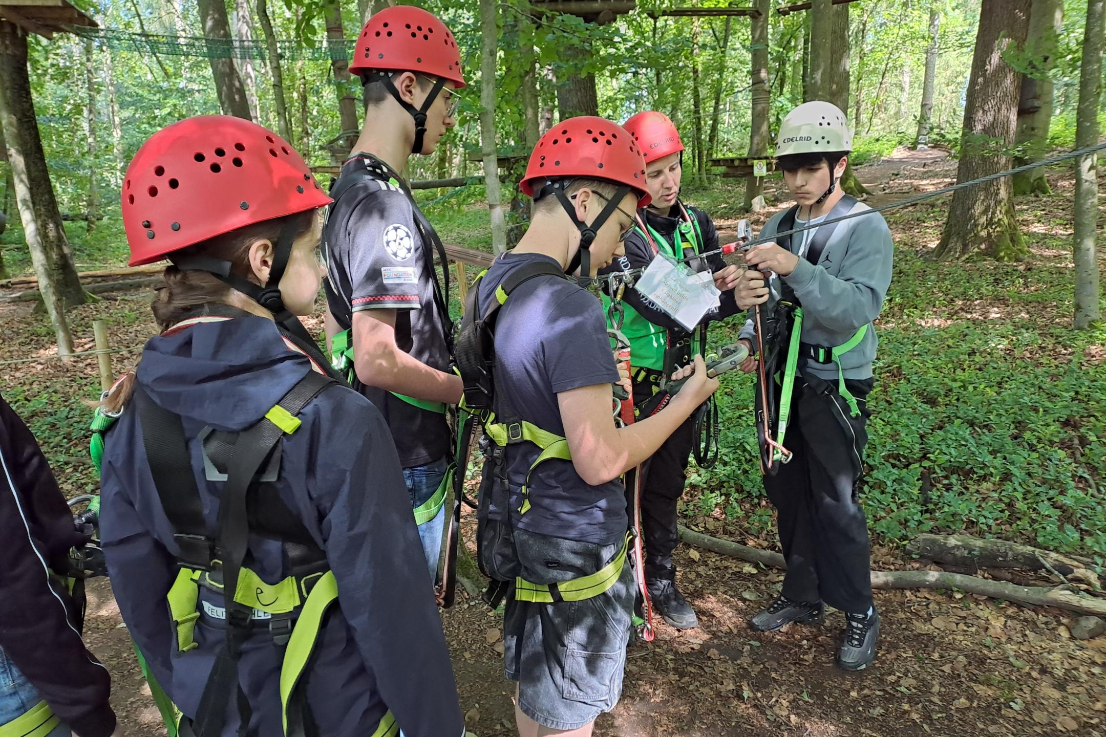 Eine Gruppe von Jugendlichen steht im Wald zusammen und überprüft gemeinsam Kletterausrüstung wie Karabiner und Sicherungsseile. Alle tragen Helme und Gurte und bereiten sich auf den Kletterparcours vor.