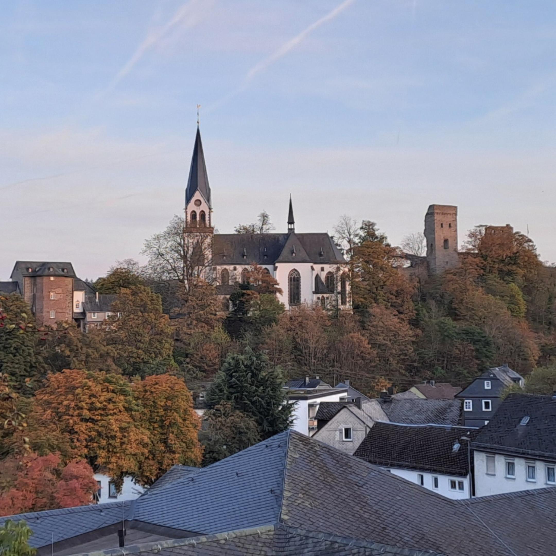 Die katholische Kirche von Kastellaun erhebt sich auf einer bewaldeten Anhöhe über dem Ort. Der helle Kirchenbau mit seinem hohen, spitzen Turm steht inmitten herbstlich gefärbter Bäume. Rechts neben der Kirche ist ein historischer Burgturm zu erkennen. Im Vordergrund liegen die Dächer mehrerer Wohnhäuser, eingerahmt von Bäumen und Sträuchern. Über der Landschaft spannt sich ein klarer blauer Himmel mit mehreren Kondensstreifen.