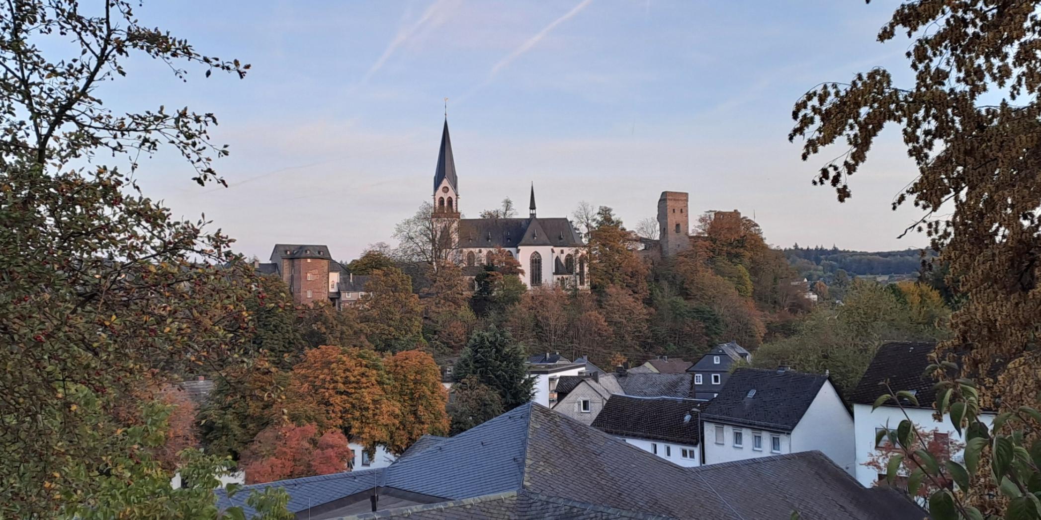 Die katholische Kirche von Kastellaun erhebt sich auf einer bewaldeten Anhöhe über dem Ort. Der helle Kirchenbau mit seinem hohen, spitzen Turm steht inmitten herbstlich gefärbter Bäume. Rechts neben der Kirche ist ein historischer Burgturm zu erkennen. Im Vordergrund liegen die Dächer mehrerer Wohnhäuser, eingerahmt von Bäumen und Sträuchern. Über der Landschaft spannt sich ein klarer blauer Himmel mit mehreren Kondensstreifen.