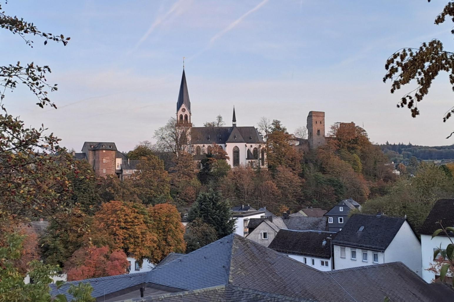 Die katholische Kirche von Kastellaun erhebt sich auf einer bewaldeten Anhöhe über dem Ort. Der helle Kirchenbau mit seinem hohen, spitzen Turm steht inmitten herbstlich gefärbter Bäume. Rechts neben der Kirche ist ein historischer Burgturm zu erkennen. Im Vordergrund liegen die Dächer mehrerer Wohnhäuser, eingerahmt von Bäumen und Sträuchern. Über der Landschaft spannt sich ein klarer blauer Himmel mit mehreren Kondensstreifen.