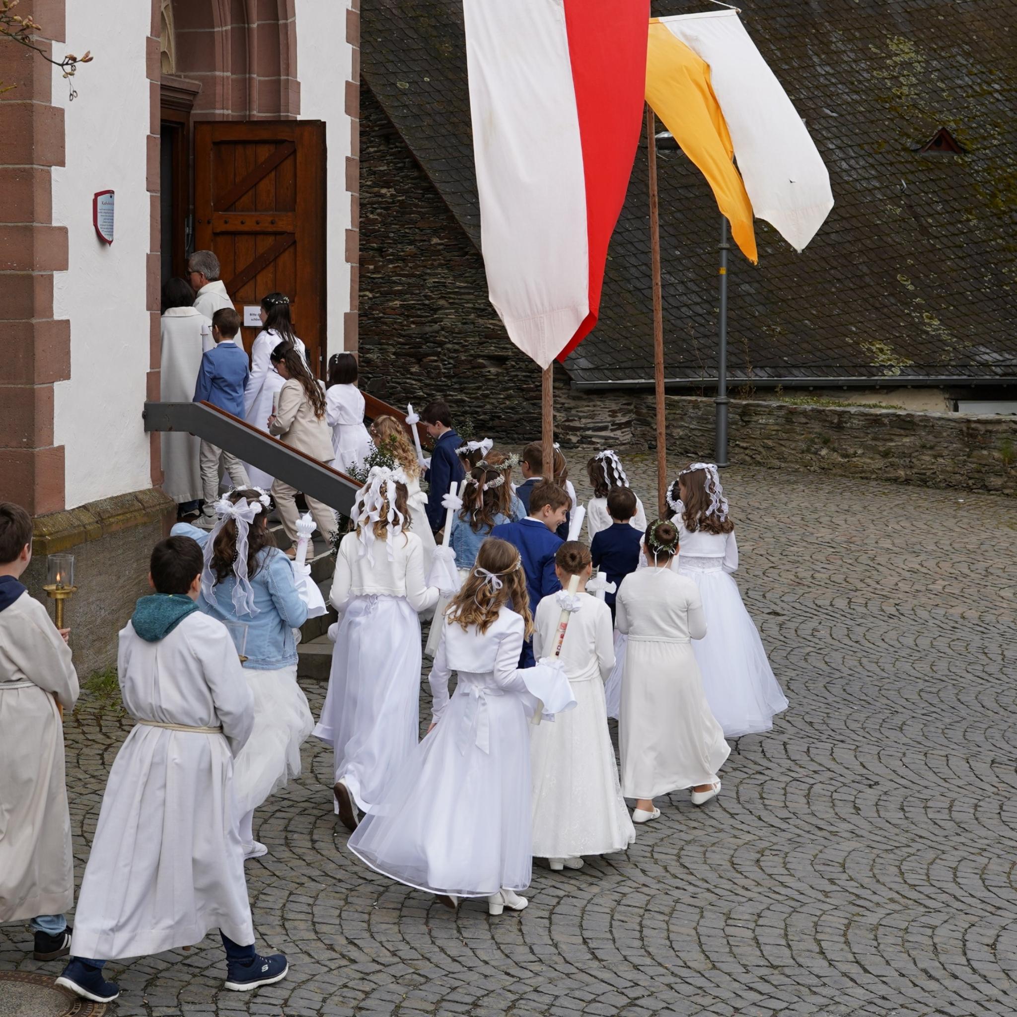 Erstkommunionkinder ziehen in die Kirche ein. Auf dem Platz stehen Fahnen.