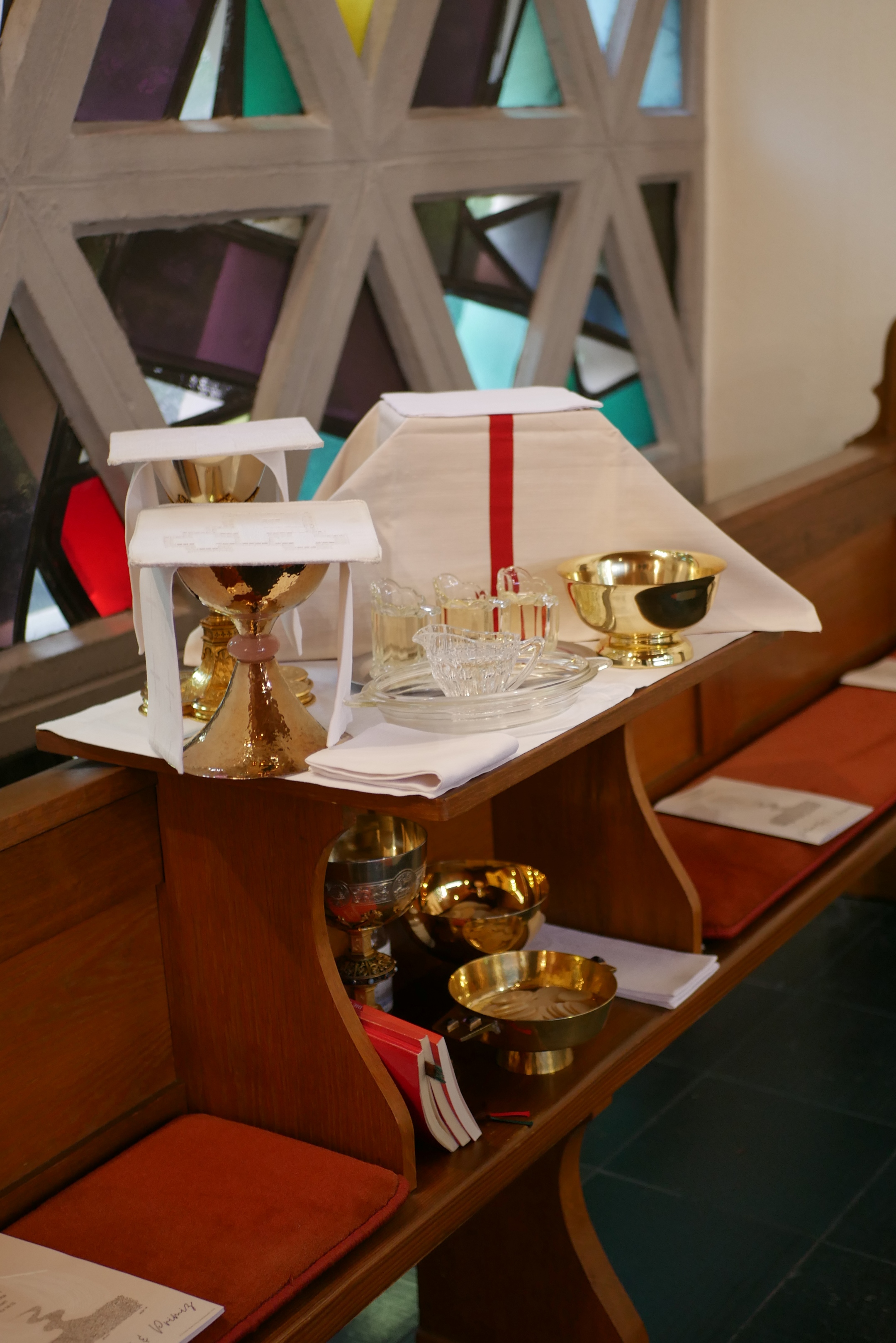 Holzbank im Kirchenraum mit darauf abgestellten liturgischen Gegenständen. Zu sehen sind mehrere goldfarbene Kelche, eine Patene, Glasgefäße, kleine Glaskännchen, gefaltete weiße Tücher und ein aufgeschlagenes Altartuch mit rotem Mittelstreifen. Im Hintergrund befindet sich ein farbiges Glasfenster mit geometrischem Muster.