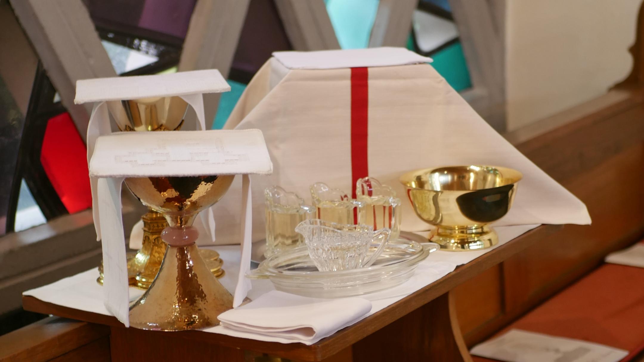 Holzbank im Kirchenraum mit darauf abgestellten liturgischen Gegenständen. Zu sehen sind mehrere goldfarbene Kelche, eine Patene, Glasgefäße, kleine Glaskännchen, gefaltete weiße Tücher und ein aufgeschlagenes Altartuch mit rotem Mittelstreifen. Im Hintergrund befindet sich ein farbiges Glasfenster mit geometrischem Muster.