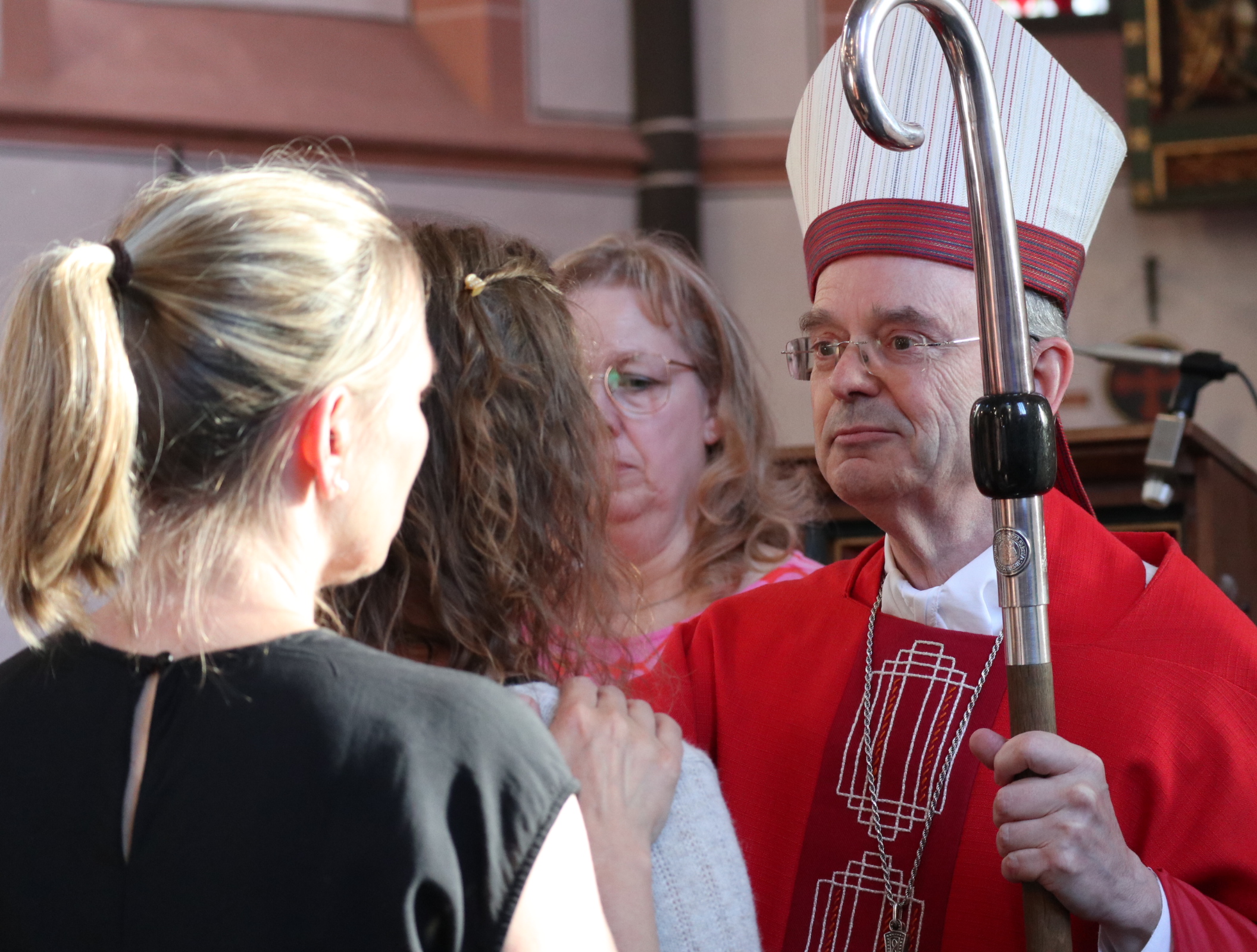 Ein katholischer Geistlicher in rotem Messgewand und mit Bischofsmütze steht in einer Kirche vor einer Gruppe von Jugendlichen. Im Vordergrund sind zwei Personen von hinten zu sehen. Der Geistliche hält einen Bischofsstab und vollzieht einen Teil der Firmfeier. Das warme Sonnenlicht fällt durch die Kirchenfenster und beleuchtet die Szene.