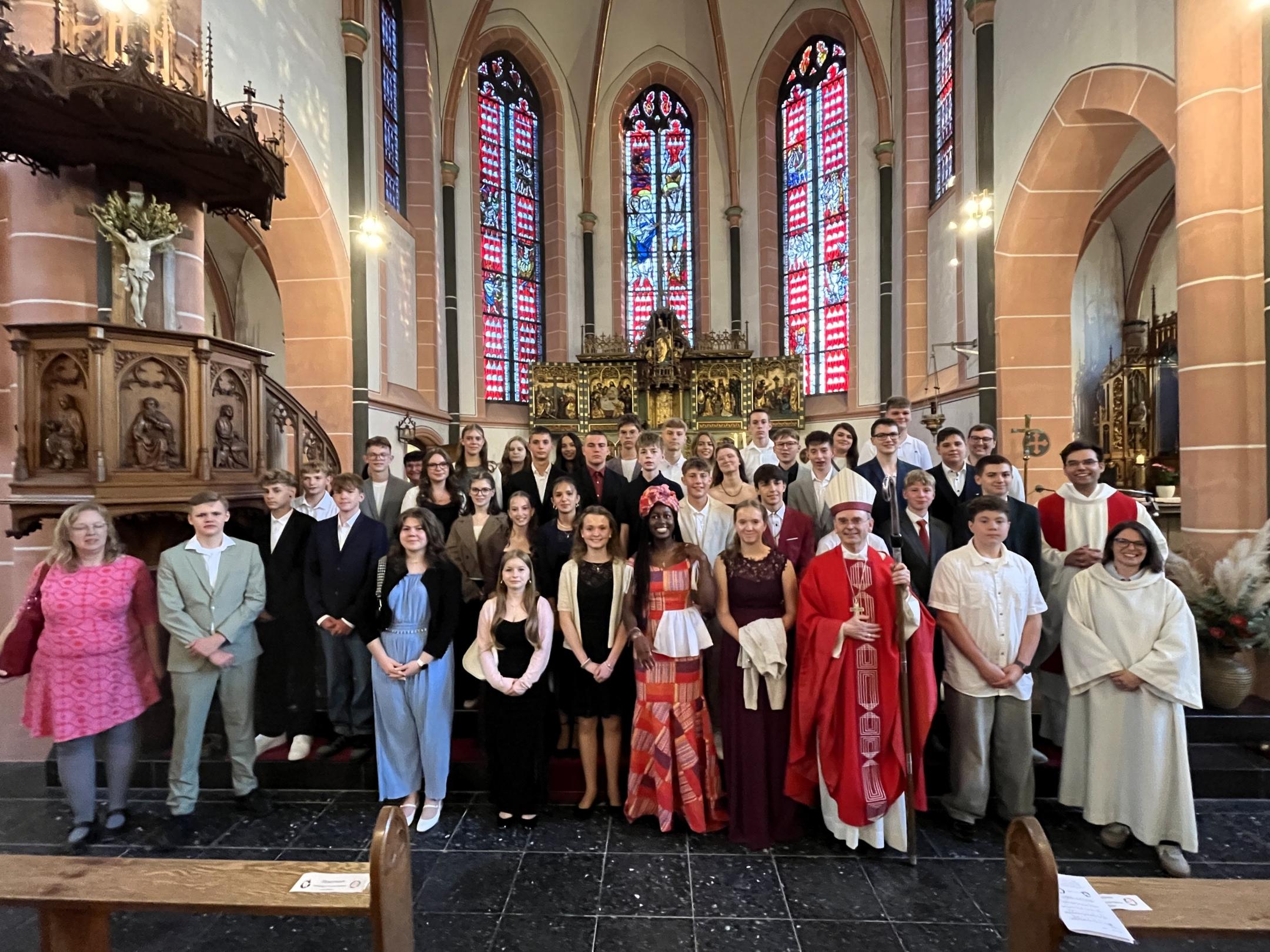 Die Firmlinge stehen gemeinsam mit Weihbischof Robert Brahm sowie weiteren Mitwirkenden vor dem Altarraum einer Kirche. Die Gruppe hat sich für ein Erinnerungsfoto auf den Stufen vor dem Altar aufgestellt. Im Hintergrund sind große farbige Kirchenfenster und historische Kirchenausstattung zu sehen.