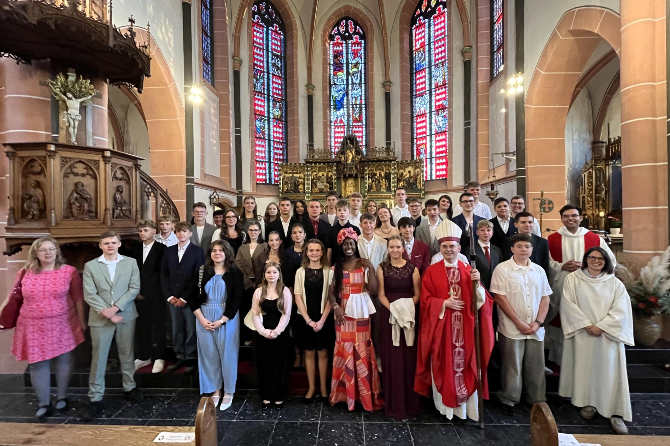 Die Firmlinge stehen gemeinsam mit Weihbischof Robert Brahm sowie weiteren Mitwirkenden vor dem Altarraum einer Kirche. Die Gruppe hat sich für ein Erinnerungsfoto auf den Stufen vor dem Altar aufgestellt. Im Hintergrund sind große farbige Kirchenfenster und historische Kirchenausstattung zu sehen.