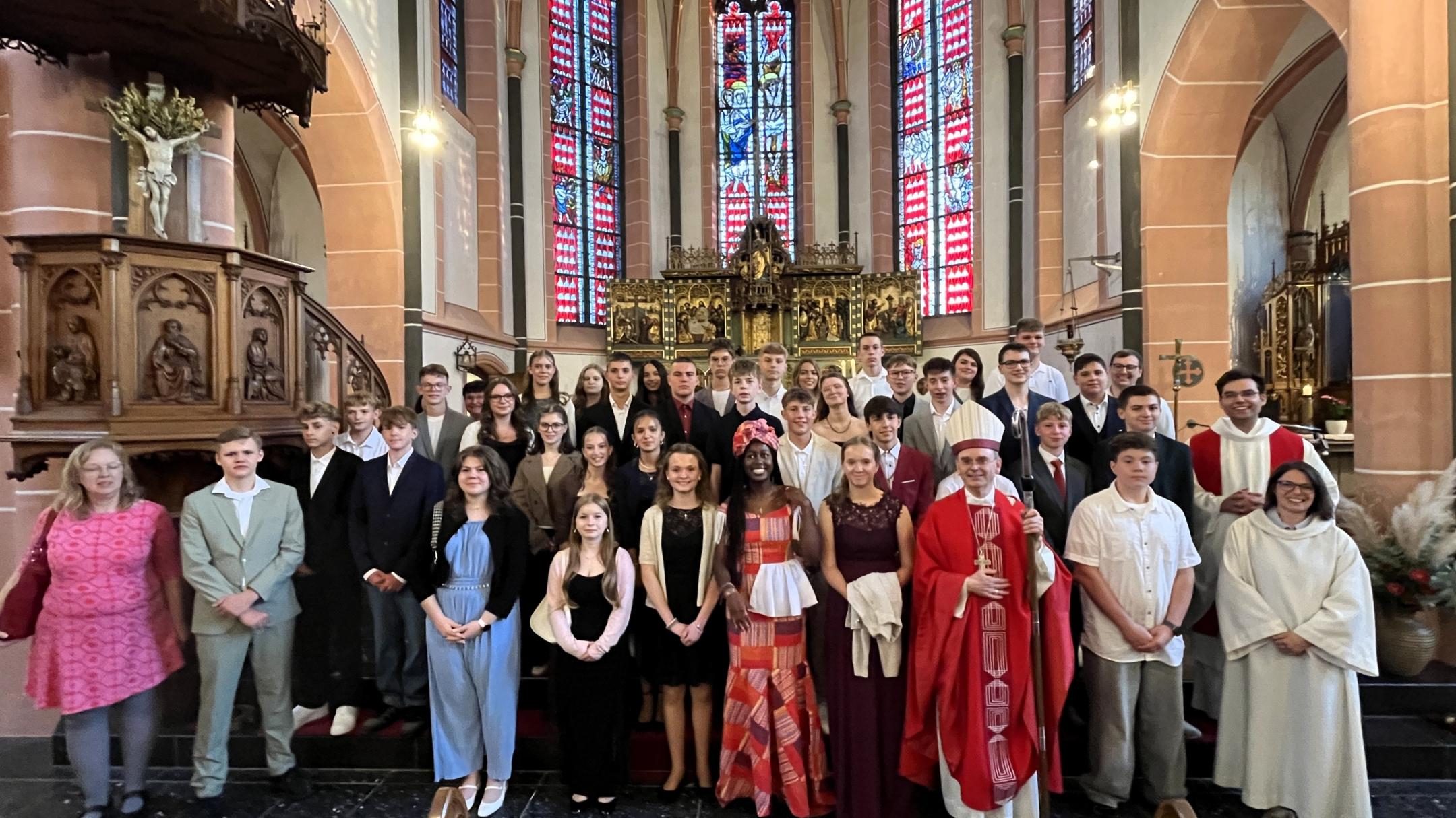Die Firmlinge stehen gemeinsam mit Weihbischof Robert Brahm sowie weiteren Mitwirkenden vor dem Altarraum einer Kirche. Die Gruppe hat sich für ein Erinnerungsfoto auf den Stufen vor dem Altar aufgestellt. Im Hintergrund sind große farbige Kirchenfenster und historische Kirchenausstattung zu sehen.