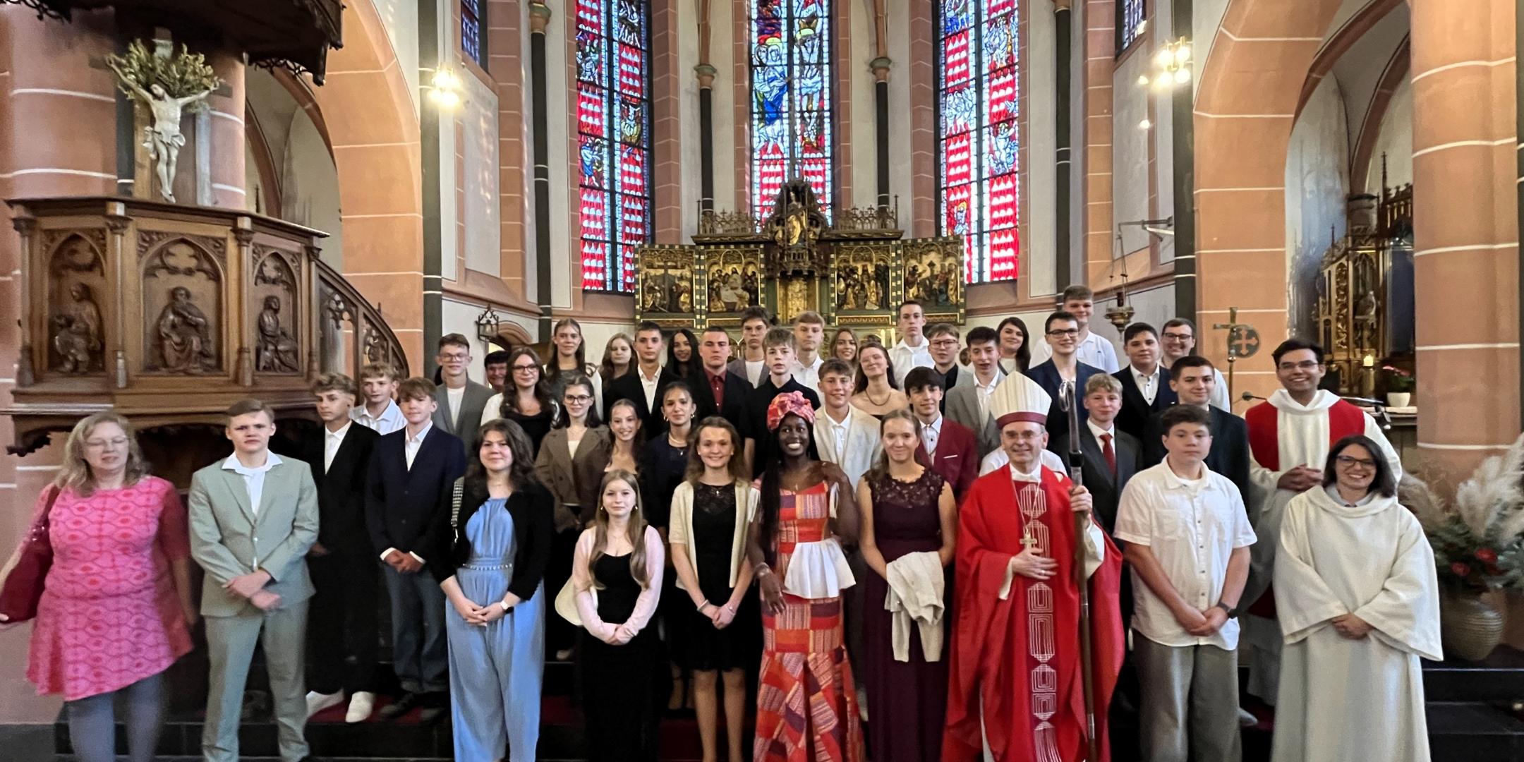 Die Firmlinge stehen gemeinsam mit Weihbischof Robert Brahm sowie weiteren Mitwirkenden vor dem Altarraum einer Kirche. Die Gruppe hat sich für ein Erinnerungsfoto auf den Stufen vor dem Altar aufgestellt. Im Hintergrund sind große farbige Kirchenfenster und historische Kirchenausstattung zu sehen.