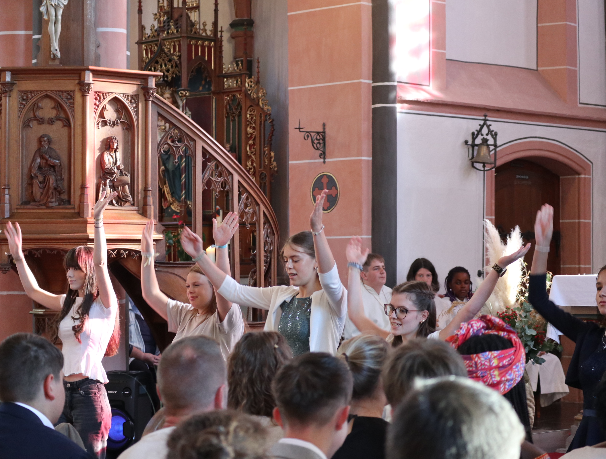 Mehrere Jugendliche führen während der Firmfeier in einer Kirche einen liturgischen Tanz auf. Mit erhobenen Armen setzen sie Gebetsgesten in Bewegung um. Die Tanzaktion findet vor den Kirchenbänken und der kunstvoll gestalteten Kanzel statt und wird von der Gemeinde verfolgt.