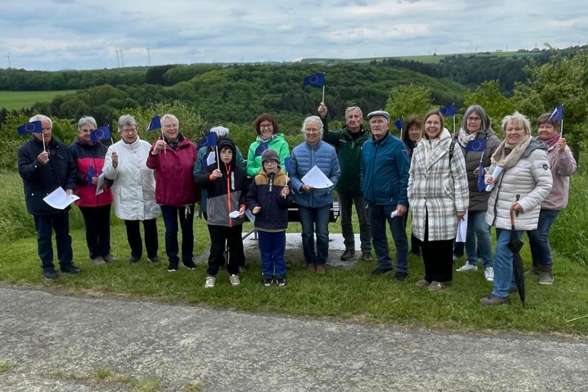 Eine Gruppe von etwa 15 Personen steht nebeneinander auf einer Wiese entlang eines Weges in einer ländlichen Umgebung. Im Hintergrund sind grüne Hügel, Bäume und mehrere Windräder zu sehen. Der Himmel ist bewölkt. Einige Personen halten kleine blaue Fahnen mit gelben Sternen (Europafahnen) in der Hand, andere halten Papierblätter. In der Mitte der Gruppe stehen auch zwei Kinder. Hinter der Gruppe befindet sich eine Bank. Die gesamte Gruppe ist nach vorne zur Kamera ausgerichtet.