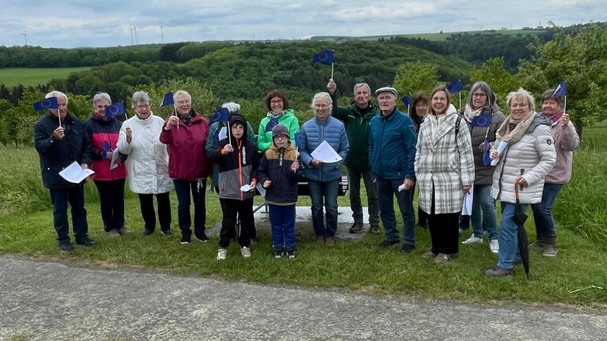 Eine Gruppe von etwa 15 Personen steht nebeneinander auf einer Wiese entlang eines Weges in einer ländlichen Umgebung. Im Hintergrund sind grüne Hügel, Bäume und mehrere Windräder zu sehen. Der Himmel ist bewölkt. Einige Personen halten kleine blaue Fahnen mit gelben Sternen (Europafahnen) in der Hand, andere halten Papierblätter. In der Mitte der Gruppe stehen auch zwei Kinder. Hinter der Gruppe befindet sich eine Bank. Die gesamte Gruppe ist nach vorne zur Kamera ausgerichtet.