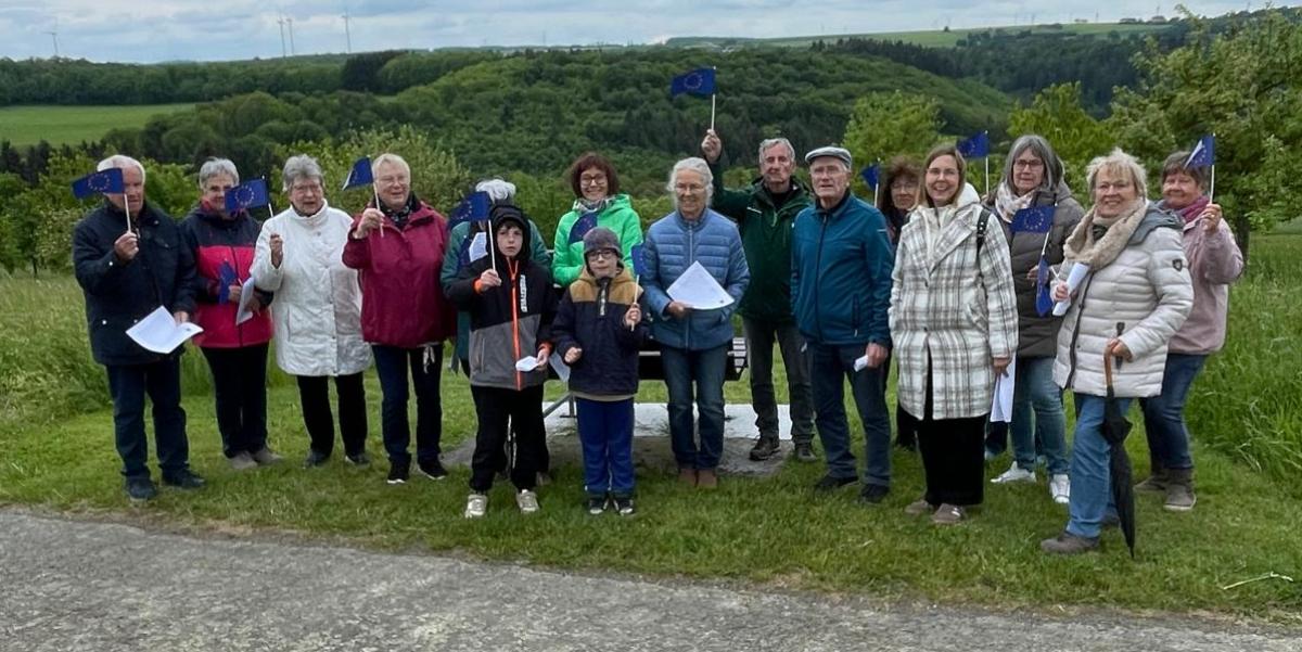 Eine Gruppe von etwa 15 Personen steht nebeneinander auf einer Wiese entlang eines Weges in einer ländlichen Umgebung. Im Hintergrund sind grüne Hügel, Bäume und mehrere Windräder zu sehen. Der Himmel ist bewölkt. Einige Personen halten kleine blaue Fahnen mit gelben Sternen (Europafahnen) in der Hand, andere halten Papierblätter. In der Mitte der Gruppe stehen auch zwei Kinder. Hinter der Gruppe befindet sich eine Bank. Die gesamte Gruppe ist nach vorne zur Kamera ausgerichtet.