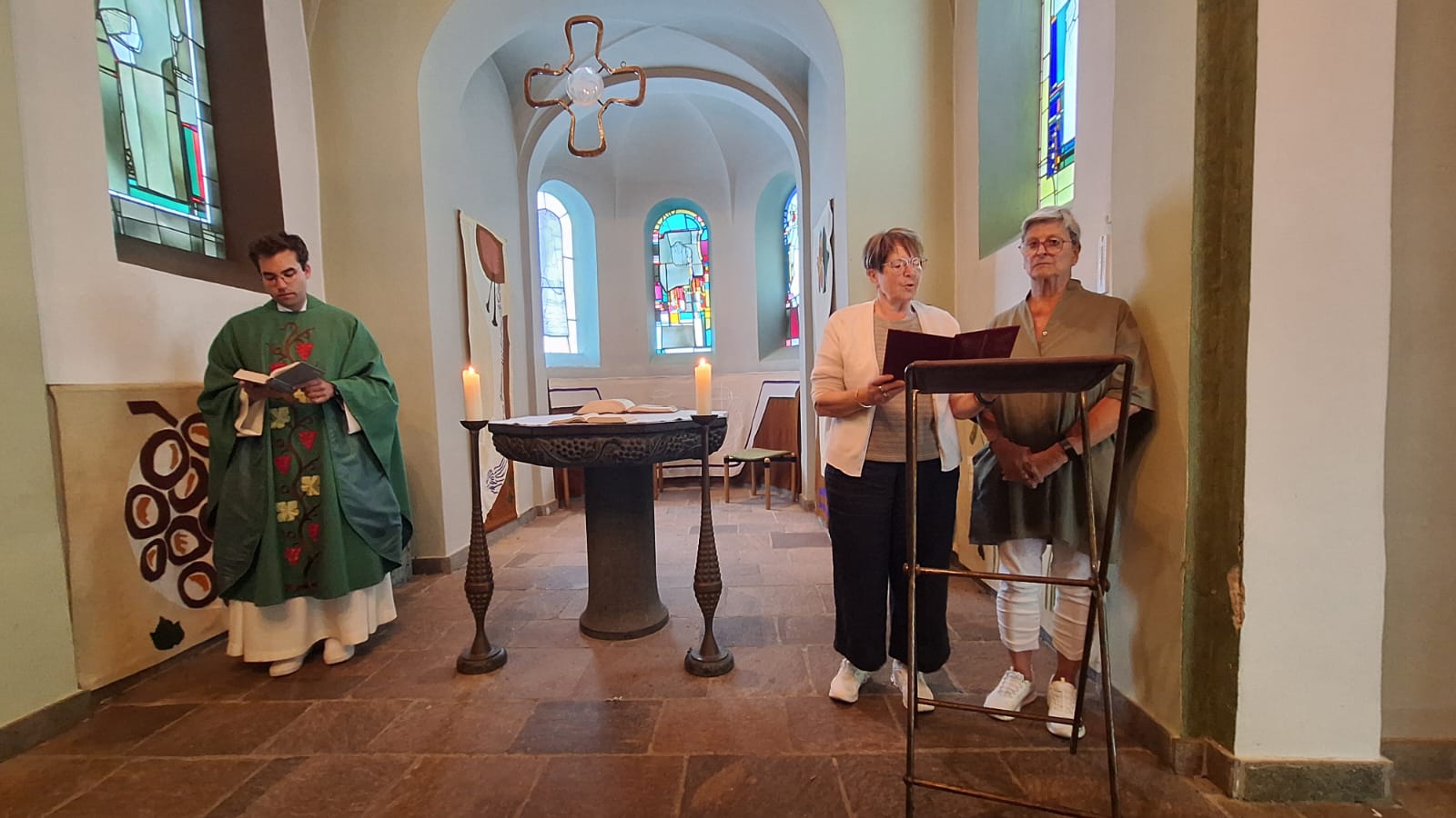 Das Bild zeigt den Innenraum einer kleinen Kapelle mit einem Altar in der Mitte. Auf dem Altar stehen zwei brennende Kerzen und ein aufgeschlagenes Buch. Links neben dem Altar steht eine Person in liturgischer Kleidung und liest aus einem Buch. Rechts neben dem Altar stehen zwei weitere Personen an einem Lesepult und tragen Texte vor. Im Hintergrund sind bunte Glasfenster zu sehen, durch die Licht in den Raum fällt. Die Architektur ist schlicht, mit hellen Wänden und Rundbögen, wodurch eine ruhige und konzentrierte Atmosphäre entsteht. (c) Gaby Mathissen Das Bild zeigt den Innenraum einer kleinen Kapelle mit einem Altar in der Mitte. Auf dem Altar stehen zwei brennende Kerzen und ein aufgeschlagenes Buch. Links neben dem Altar steht eine Person in liturgischer Kleidung und liest aus einem Buch. Rechts neben dem Altar stehen zwei weitere Personen an einem Lesepult und tragen Texte vor. Im Hintergrund sind bunte Glasfenster zu sehen, durch die Licht in den Raum fällt. Die Architektur ist schlicht, mit hellen Wänden und Rundbögen, wodurch eine ruhige und konzentrierte Atmosphäre entsteht.