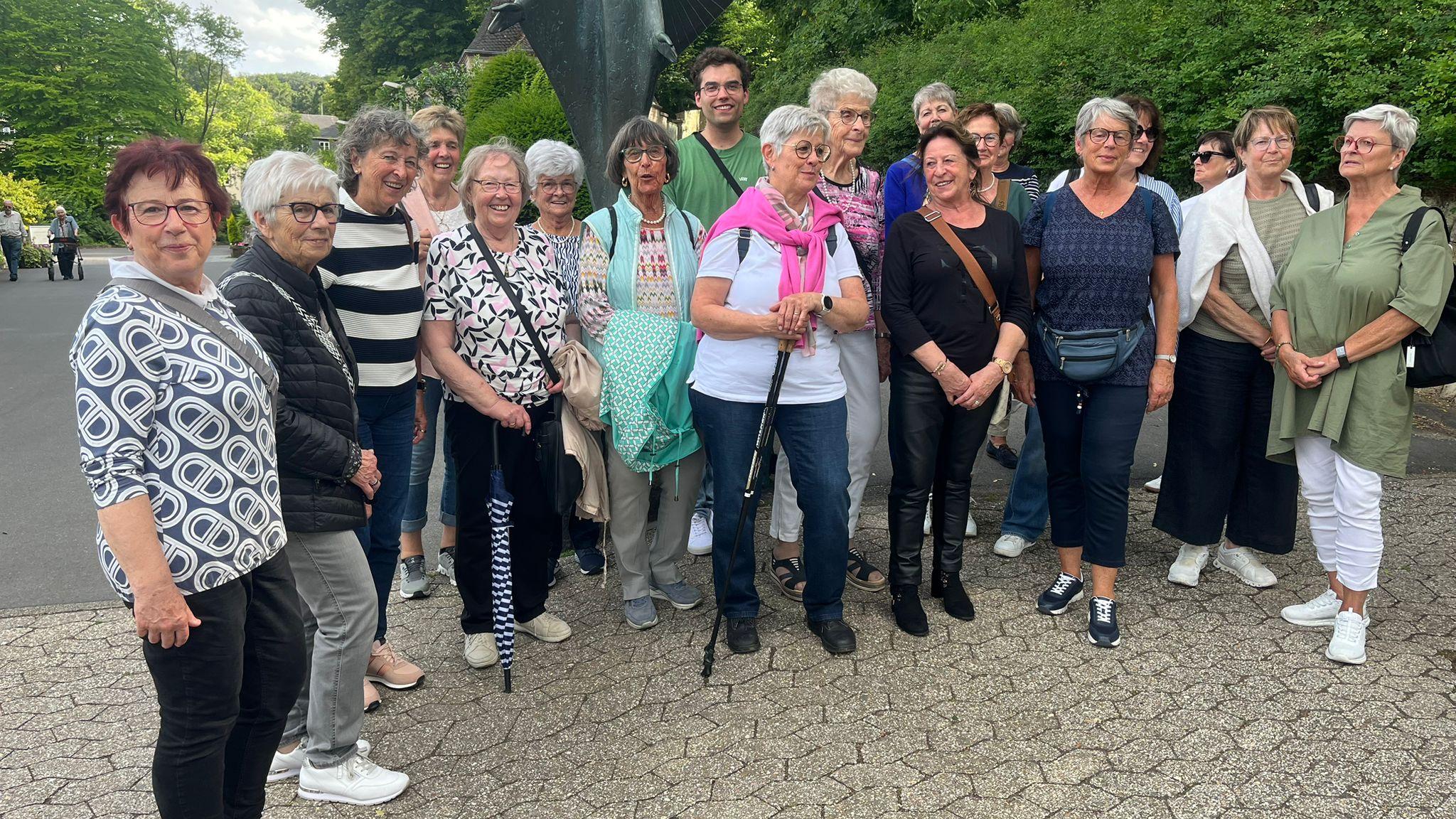 Eine größere Gruppe von Personen steht im Freien auf einem gepflasterten Weg zusammen. Hinter der Gruppe befindet sich eine große, dunkel gefärbte Skulptur mit ausgebreiteten Formen. Die Gruppe ist dicht nebeneinander aufgestellt und blickt in Richtung Kamera. Im Hintergrund sind Bäume und Grünflächen zu sehen, die den Ort als Teil des Klostergeländes erkennen lassen. Die Szene wirkt wie ein gemeinsames Erinnerungsfoto während des Ausflugs.