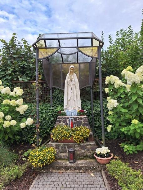 Statue der Muttergottes von Fatima in einer offenen Grotte aus Metall und Glas. Die helle Marienfigur steht auf einem gemauerten Sockel mit Blumen. Umgeben ist die Grotte von blühenden Hortensien, grünen Sträuchern und einem gepflasterten Zugang.