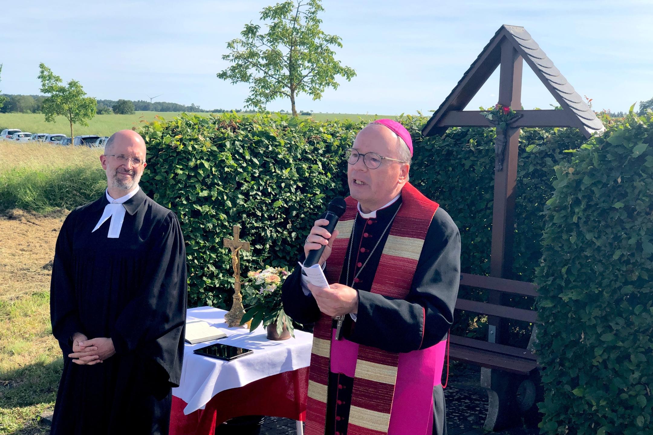 evangelischer und katholischer Geistlicher im Gespräch vor einem Kreuz und einer Hecke